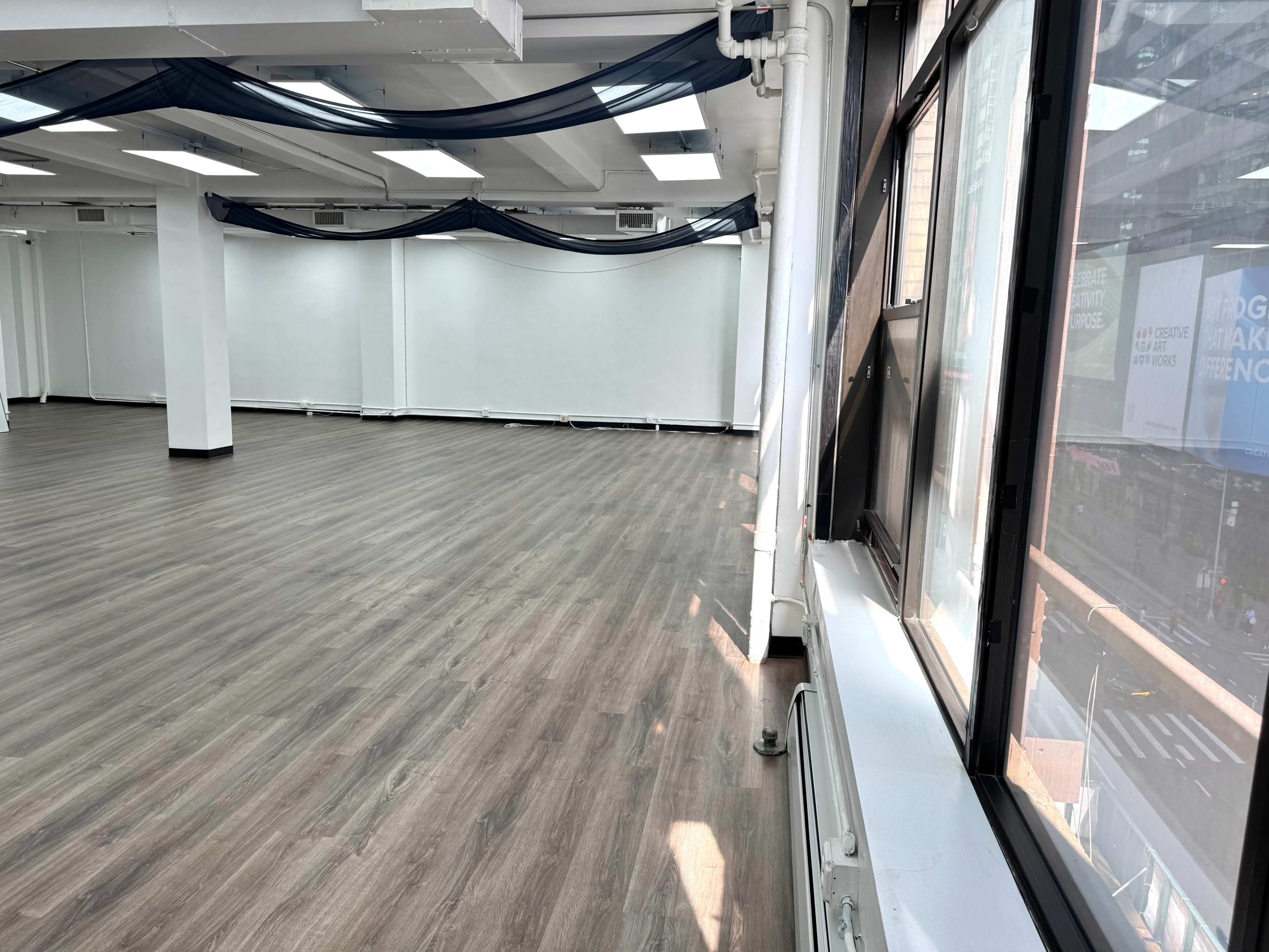 The image shows a spacious, empty room with hardwood flooring and large windows allowing natural light to enter.