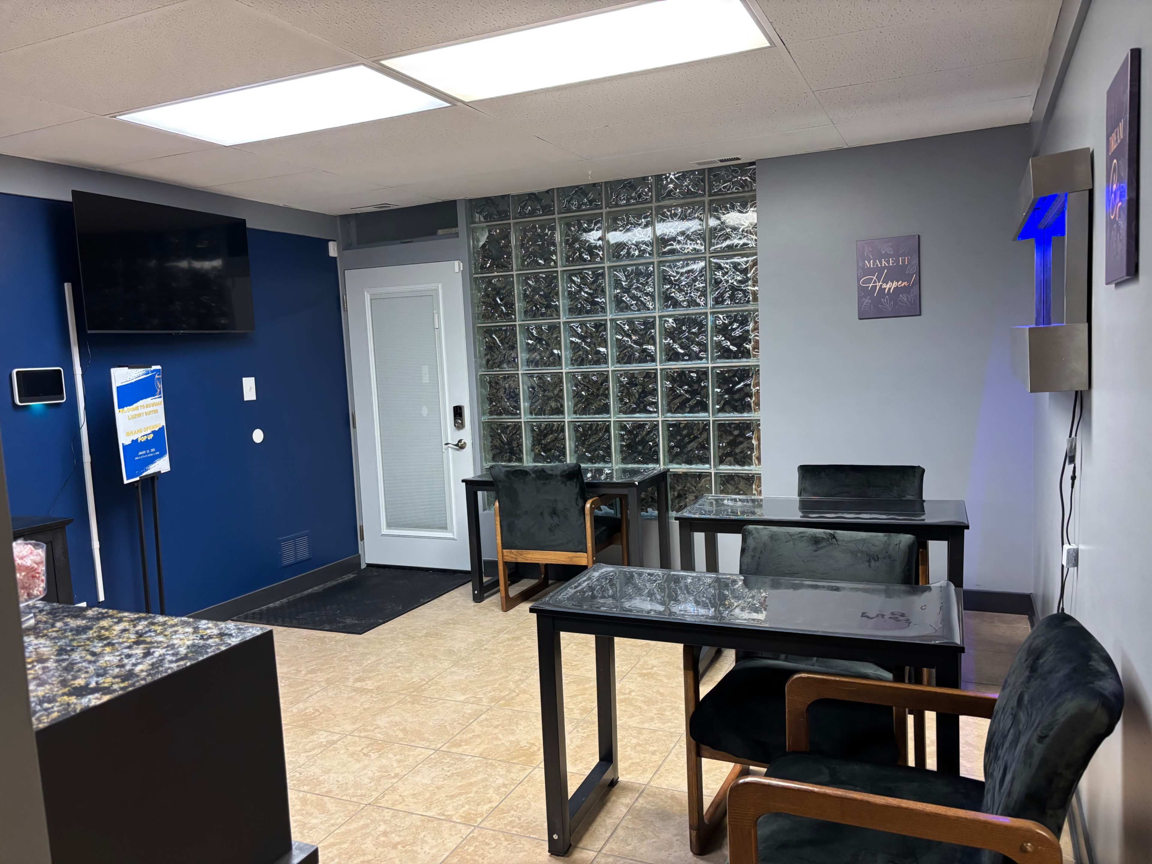 The image shows a modern waiting area with a dark blue accent wall, glass block dividers, and two black tables with chairs arranged around them.