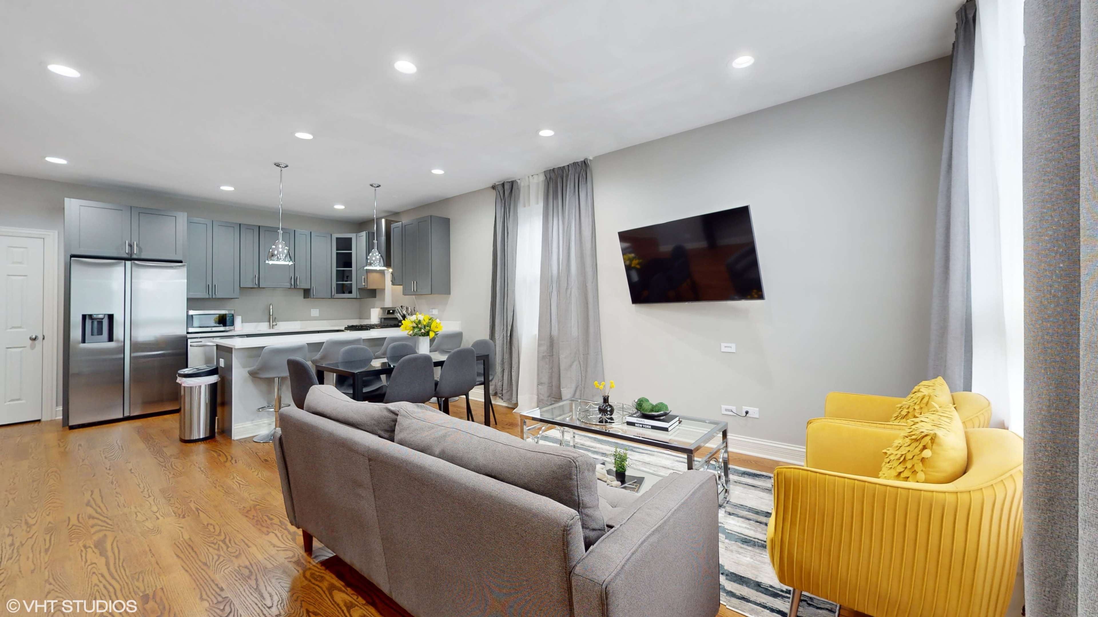 A modern living room with a gray sofa, yellow chairs, a glass coffee table, and a mounted television, adjacent to a kitchen space with gray cabinetry and a dining table.