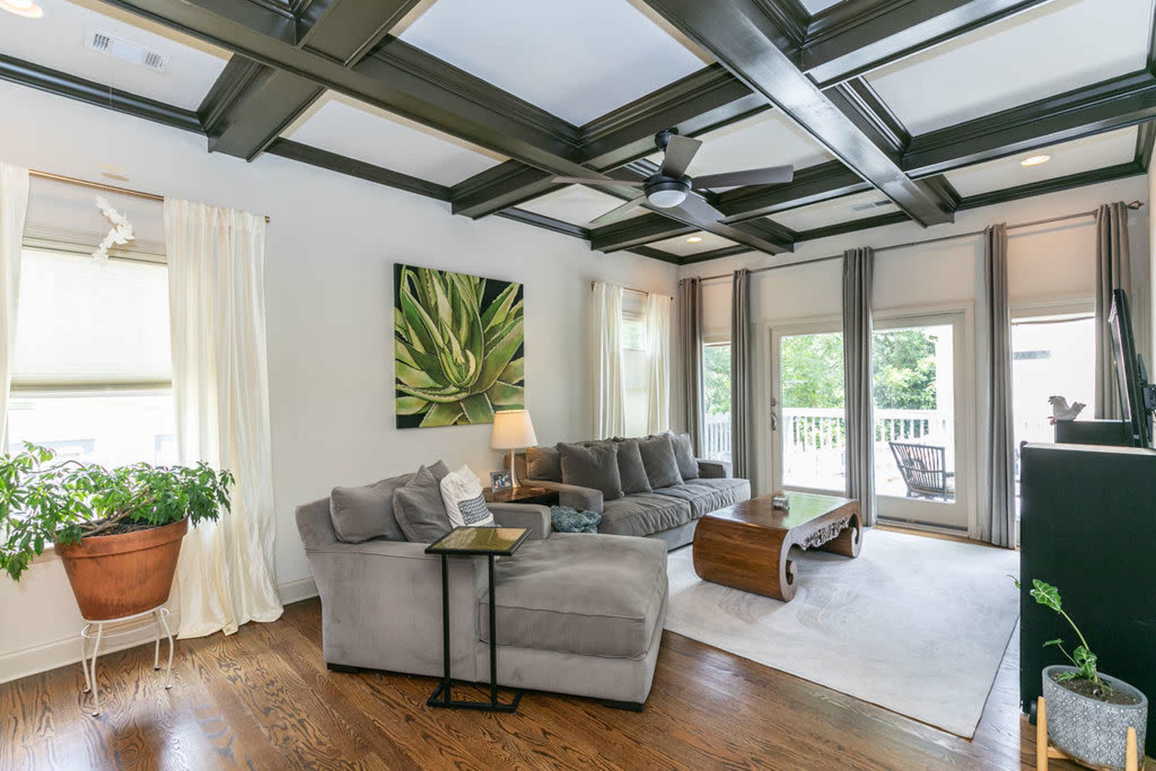 A living room features a coffered ceiling, a grey sectional sofa, a wooden coffee table, large windows, and a potted plant.