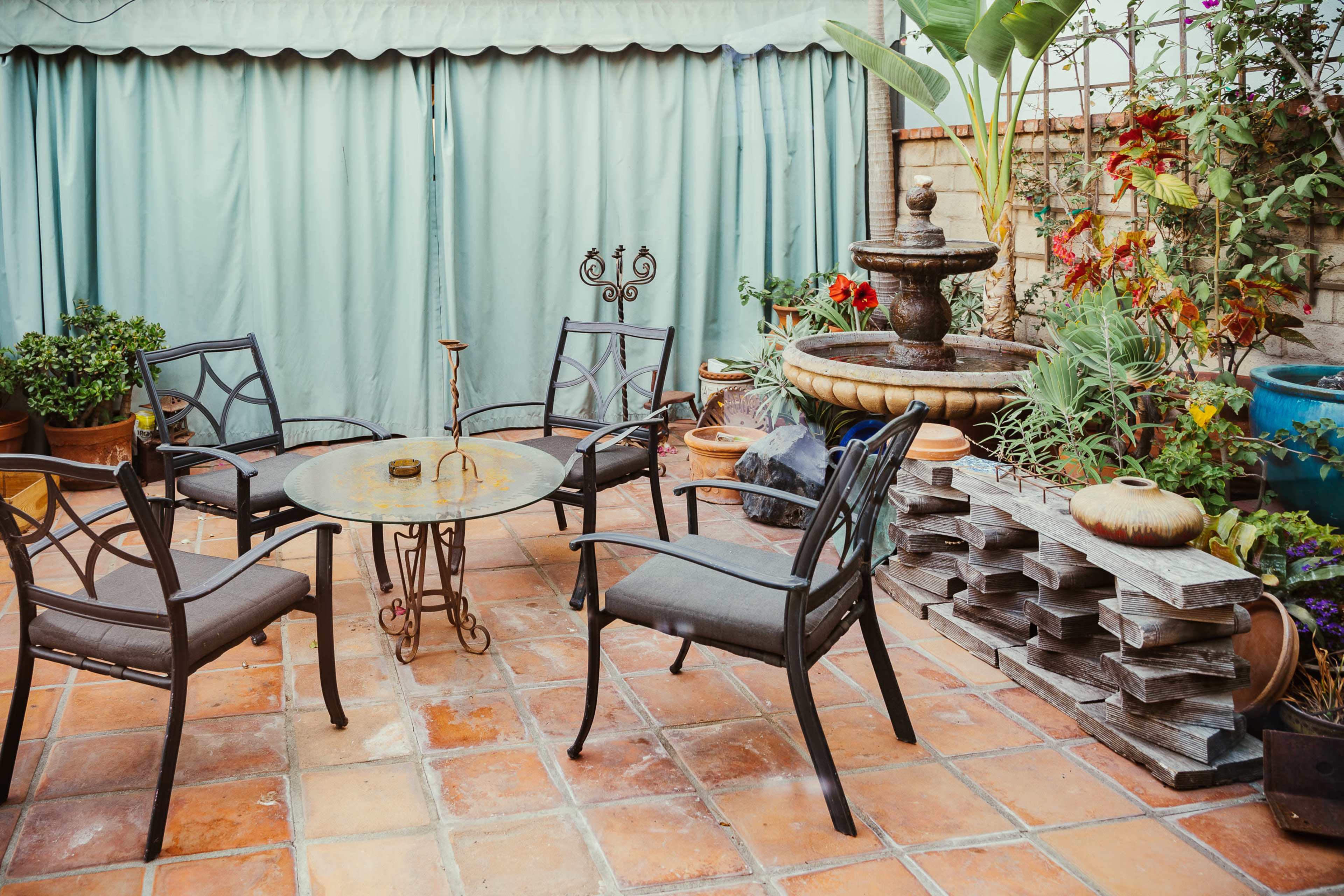 A small courtyard features a circular metal table surrounded by four chairs, a fountain, and various potted plants.