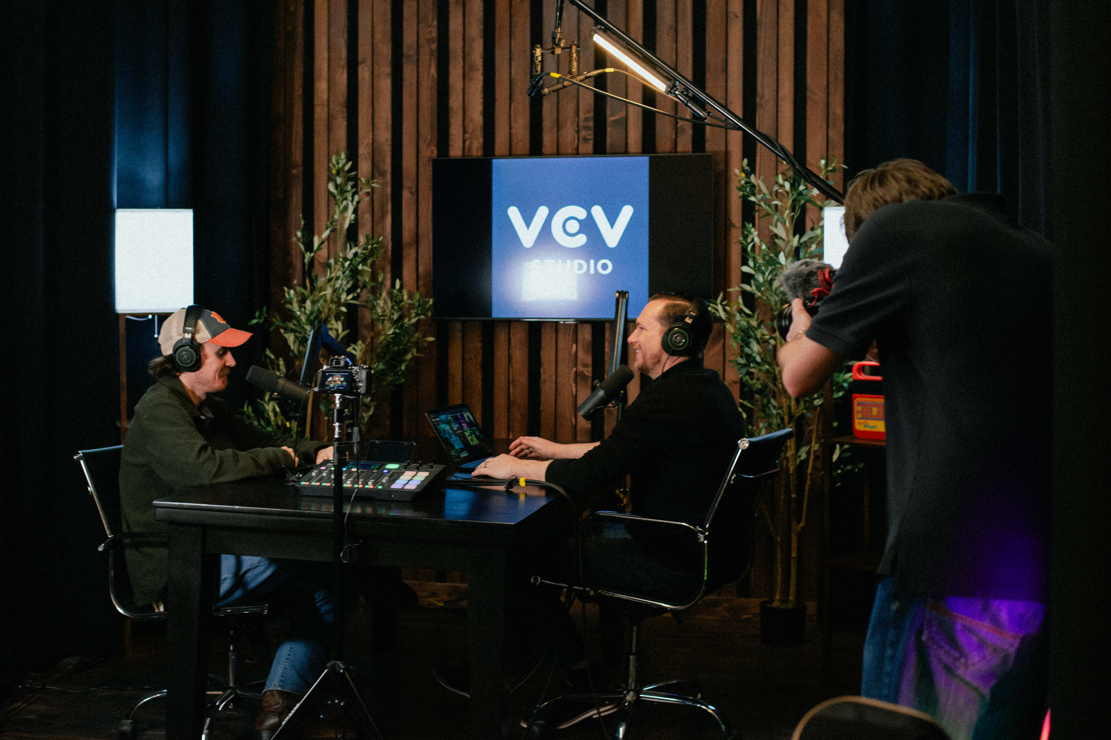Two men are seated at a desk in a studio setting, engaged in a podcast recording while a crew member operates a camera nearby.