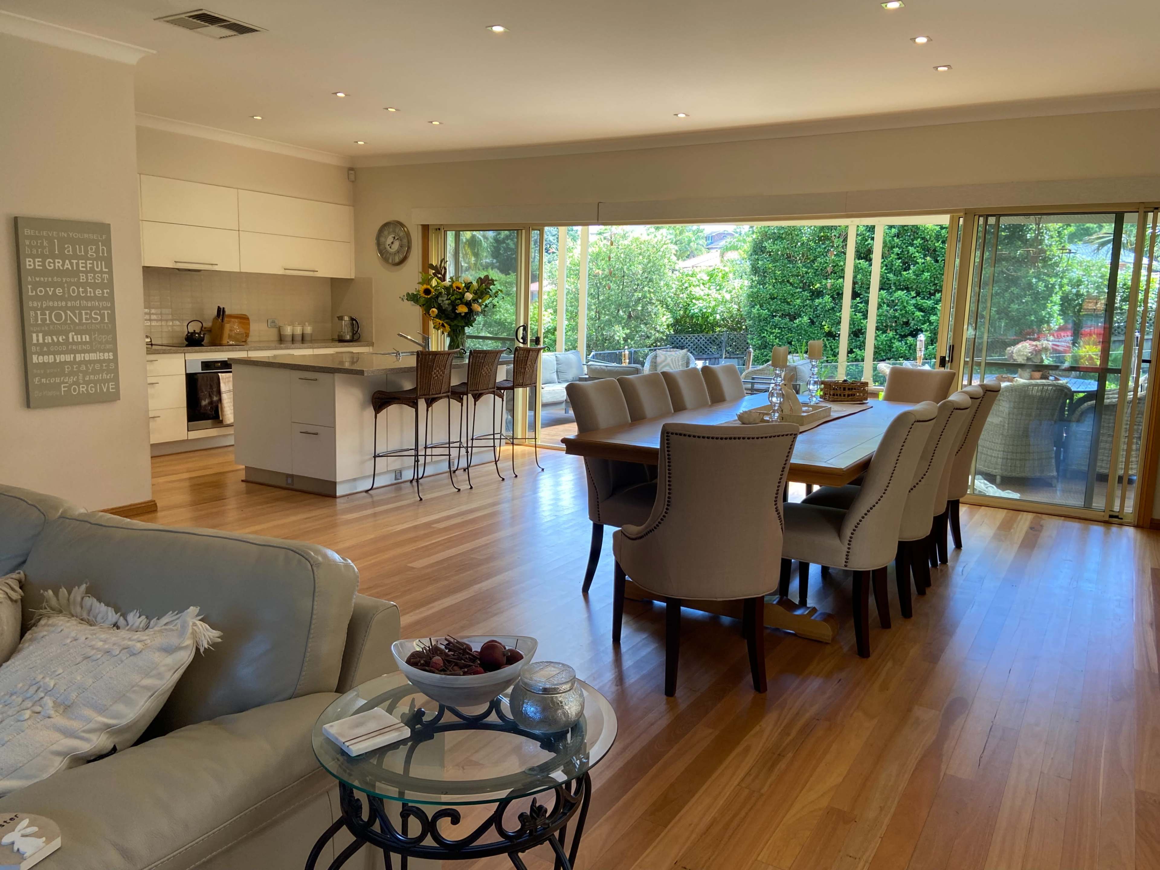 The image shows a modern, open-plan living and dining area with a kitchen, wooden floors, a large dining table, and sliding glass doors leading to an outdoor space.