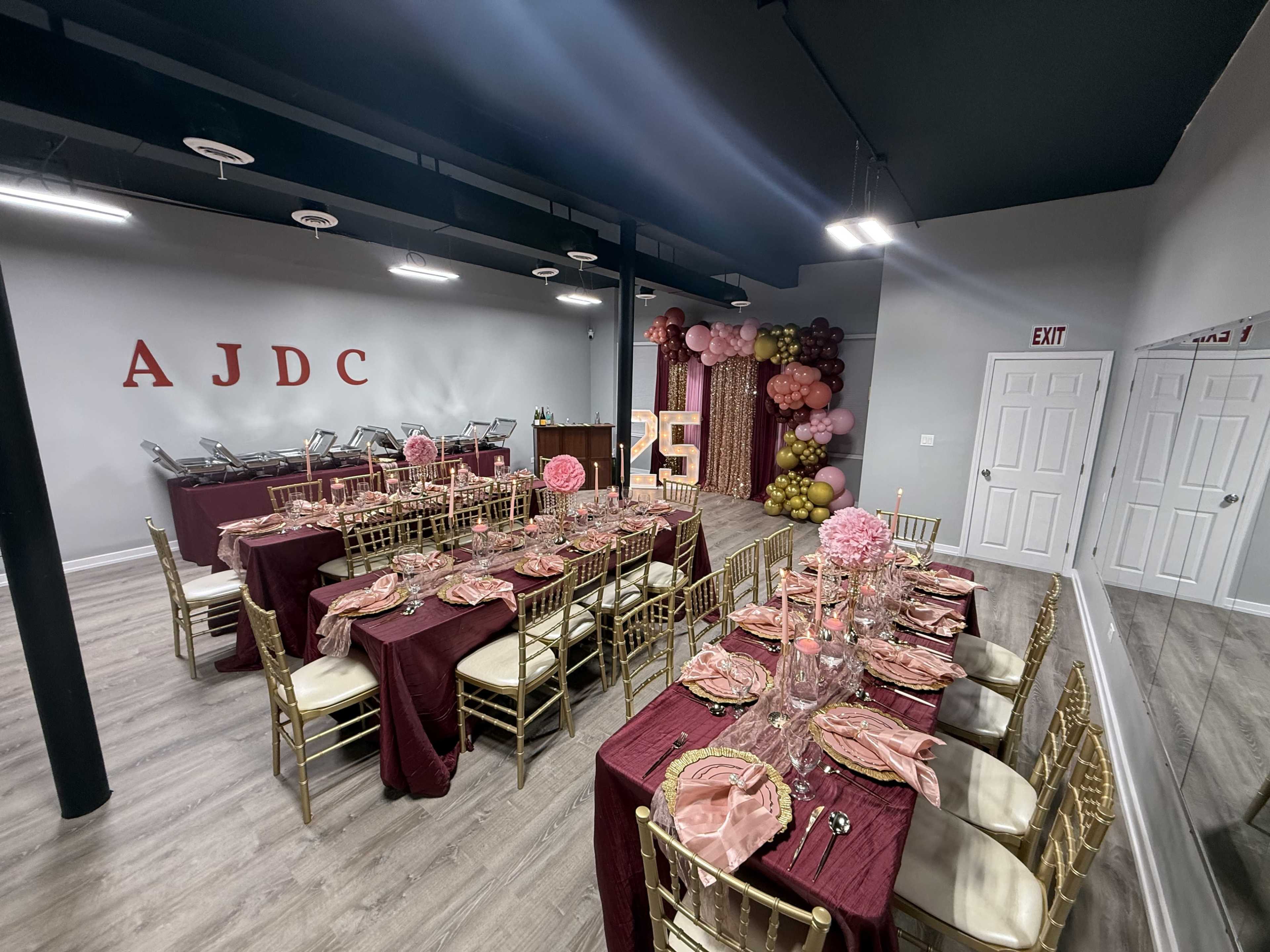 The image shows a decorated event space with tables set for a celebration, featuring burgundy tablecloths, gold chairs, and a backdrop of balloons.