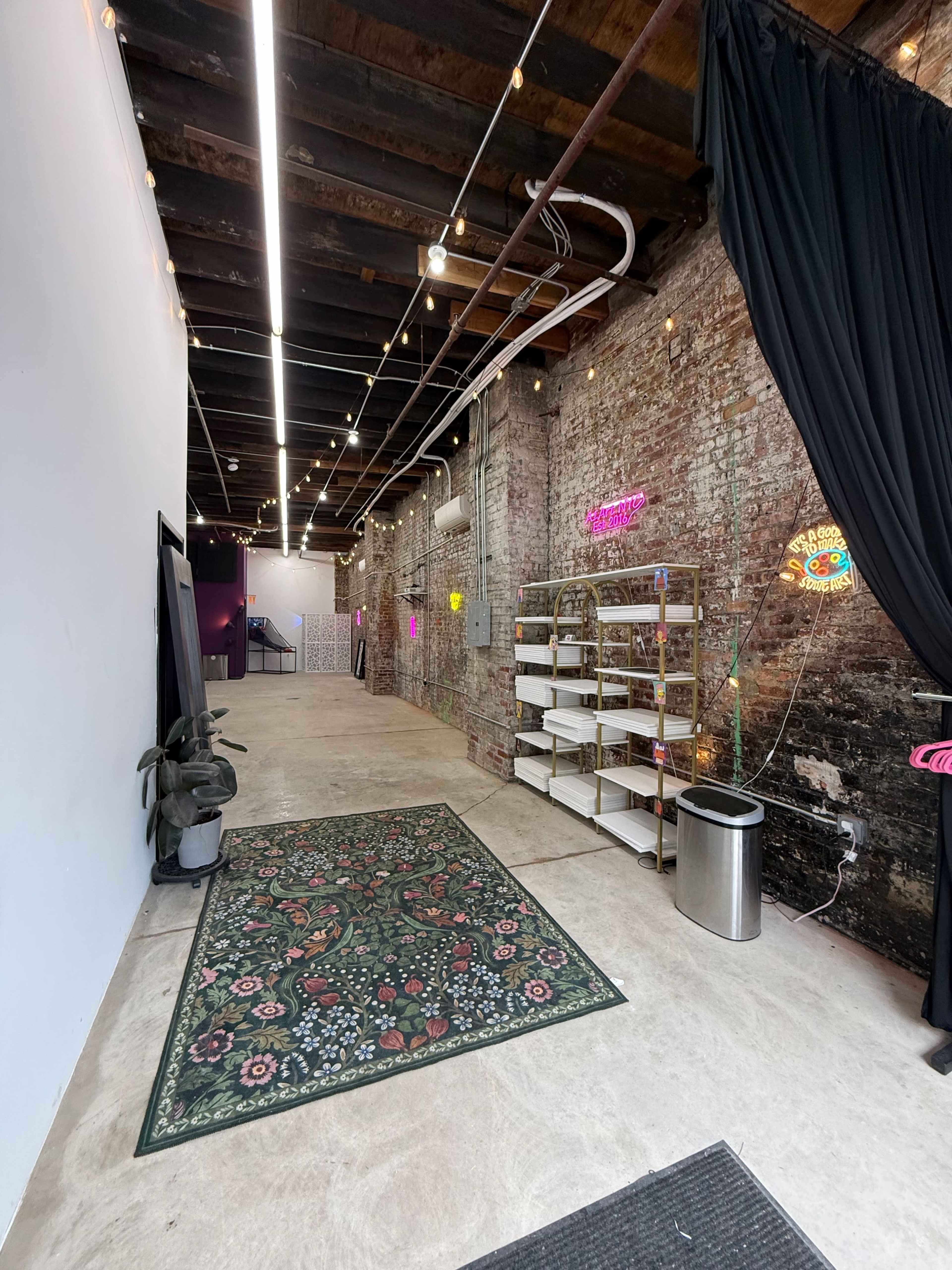 The image shows a spacious interior hallway with exposed brick walls, overhead lights, and a black curtain on the left side, leading to a brightly lit area in the background.