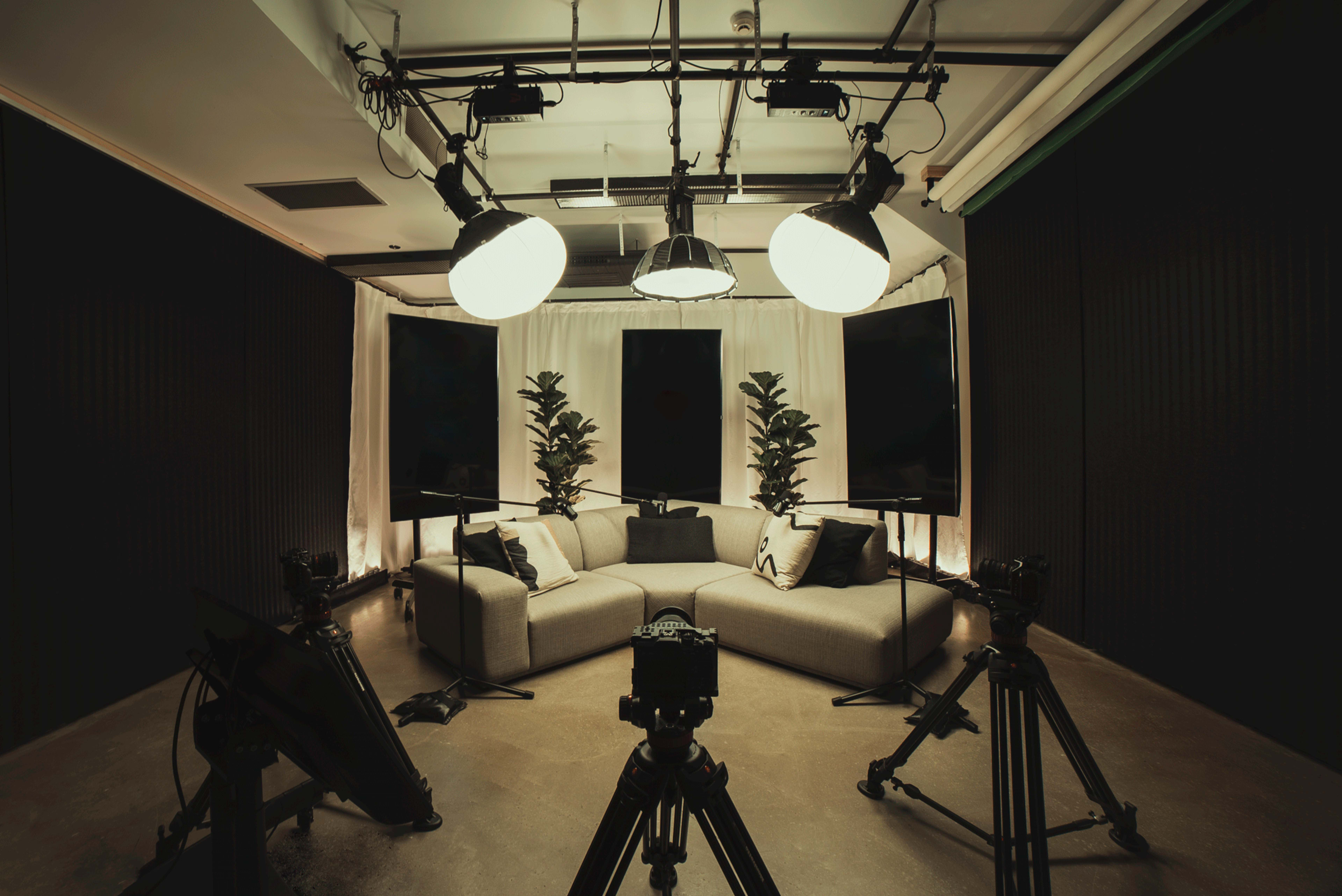 The image shows a well-lit studio setup with a gray sofa, two plants, and multiple cameras positioned around the seating area.