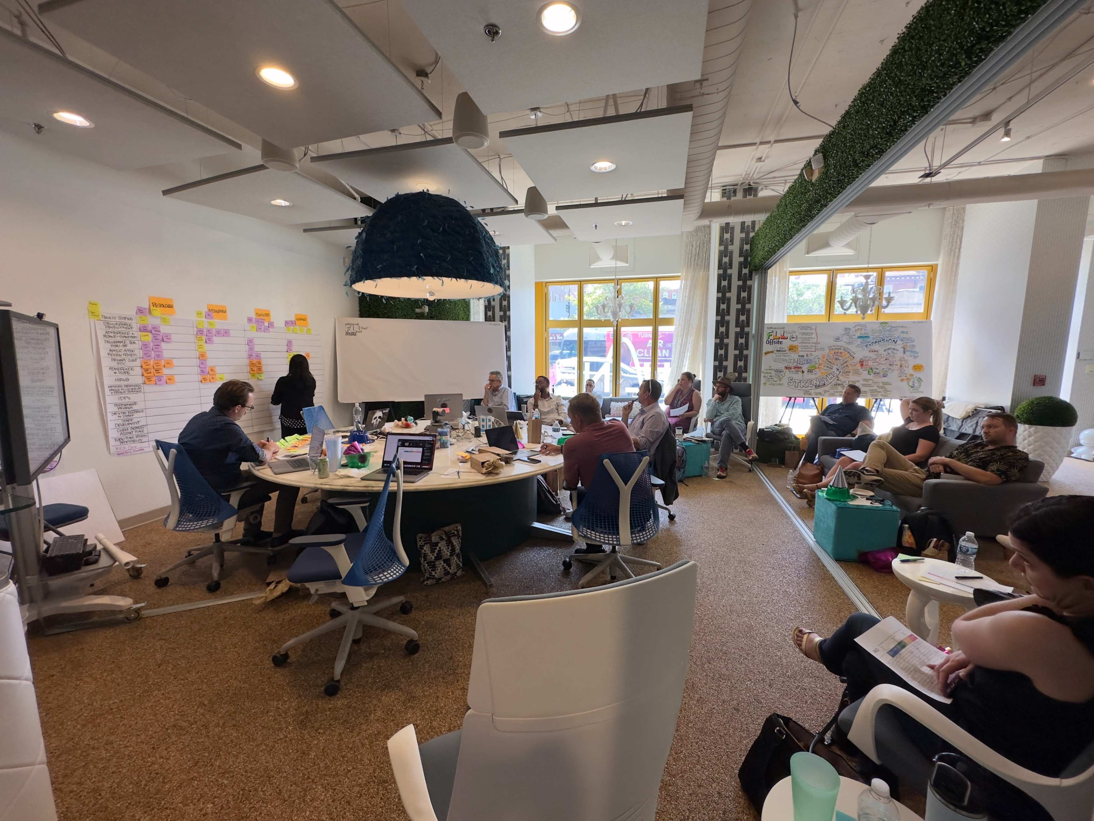 A group of people is engaging in a collaborative meeting in a brightly lit room with modern decor and whiteboards filled with notes.