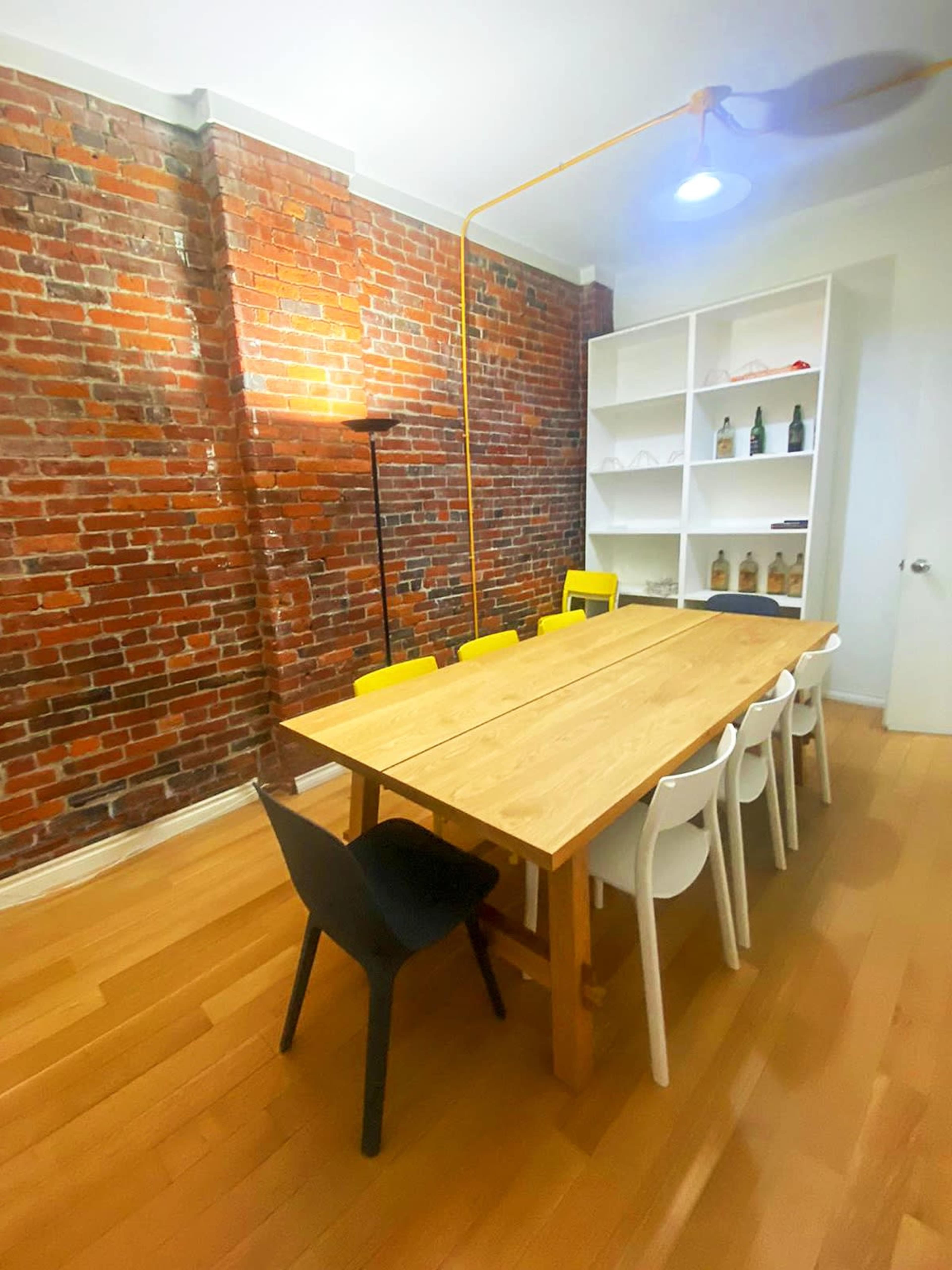A dining area featuring a long wooden table surrounded by chairs in black, white, and yellow, with a brick wall and a light fixture overhead.