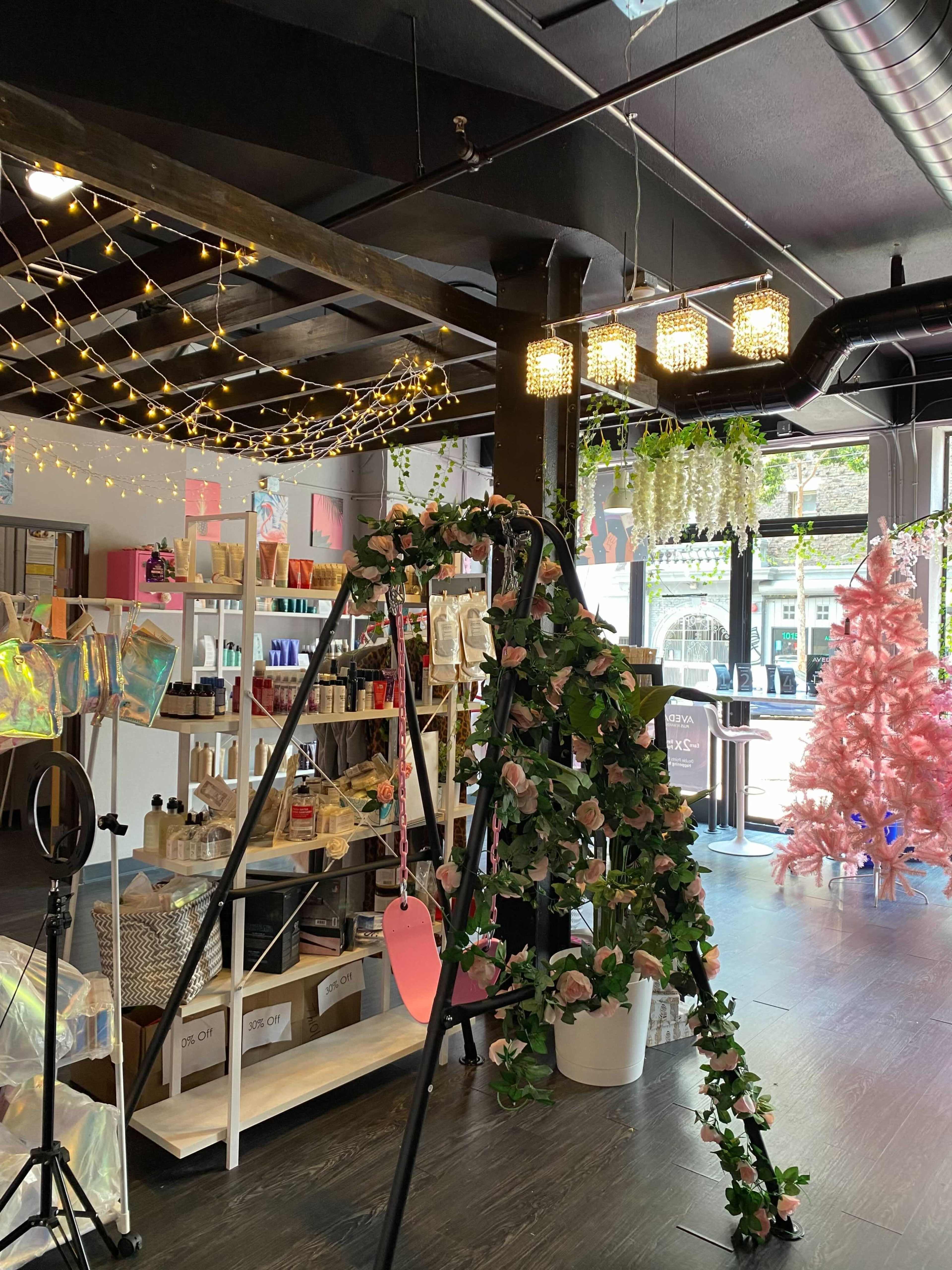 A swing decorated with artificial flowers is placed inside a retail space featuring shelves of various products and a pink Christmas tree.