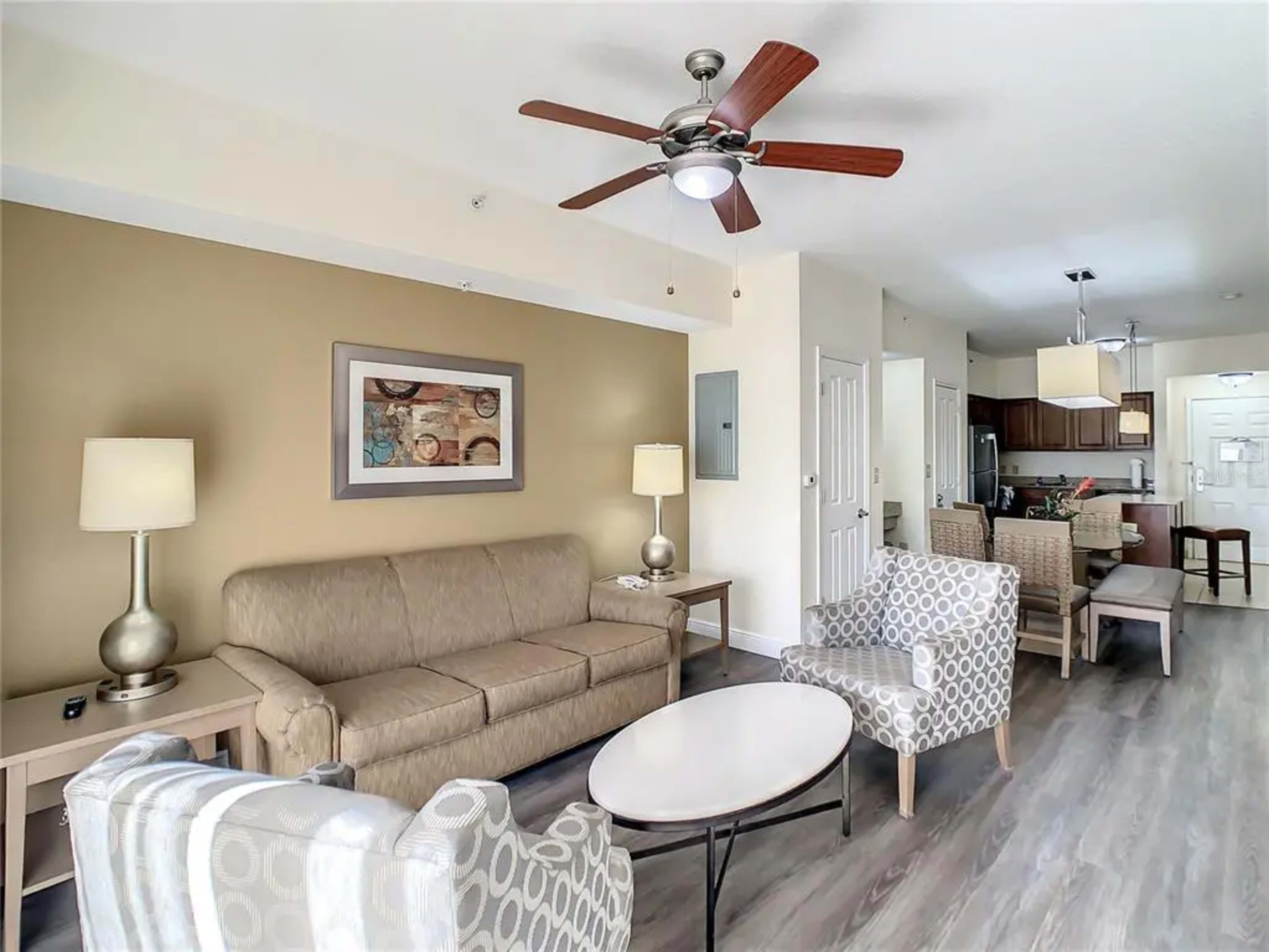 A living room with a beige sofa, two patterned chairs, a round coffee table, and a ceiling fan, leading into a kitchen area with wooden cabinets.