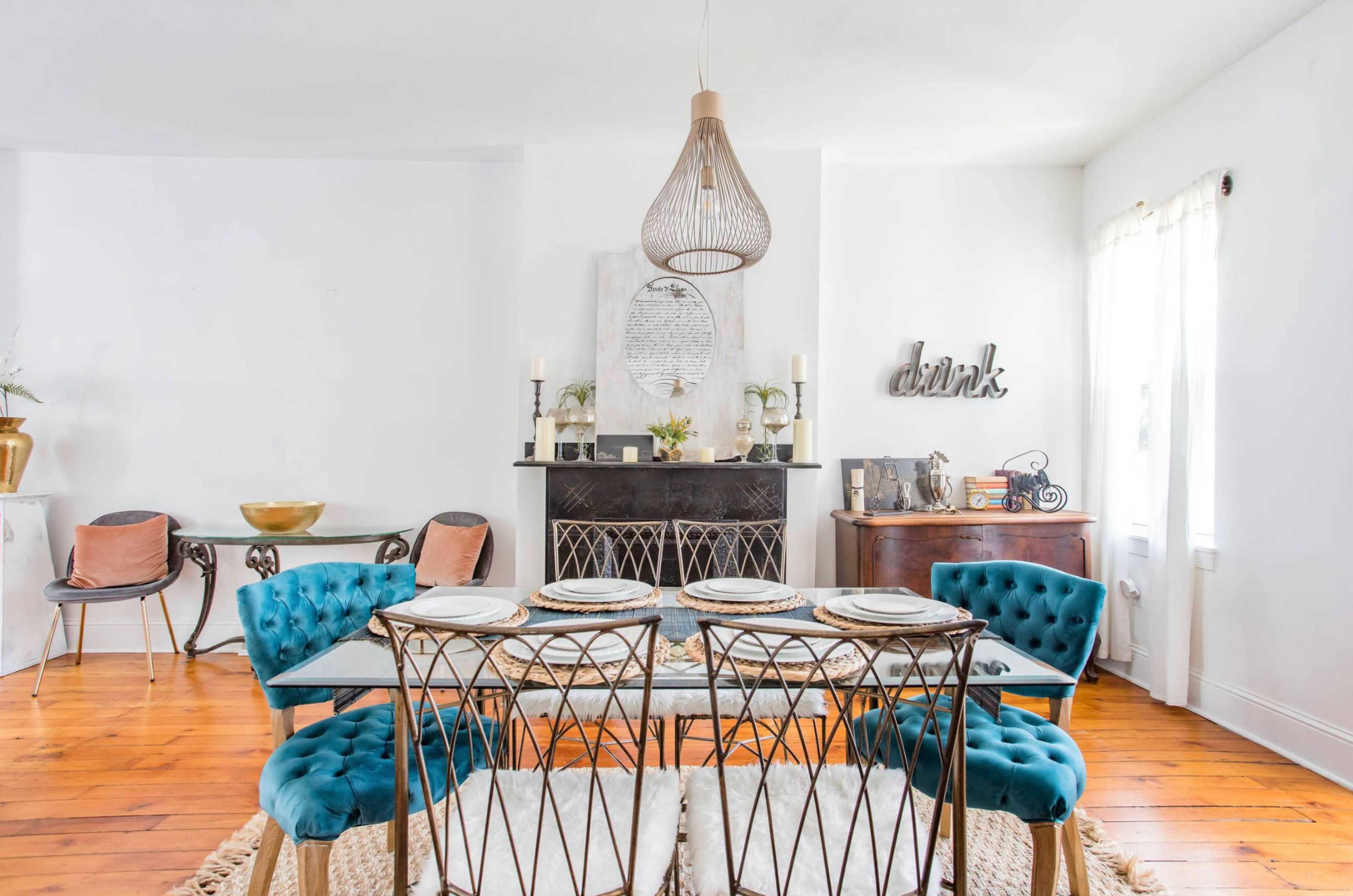 A modern dining room features a glass-top table with four teal upholstered chairs, a decorative mirror on the wall, and a fireplace adorned with candles.