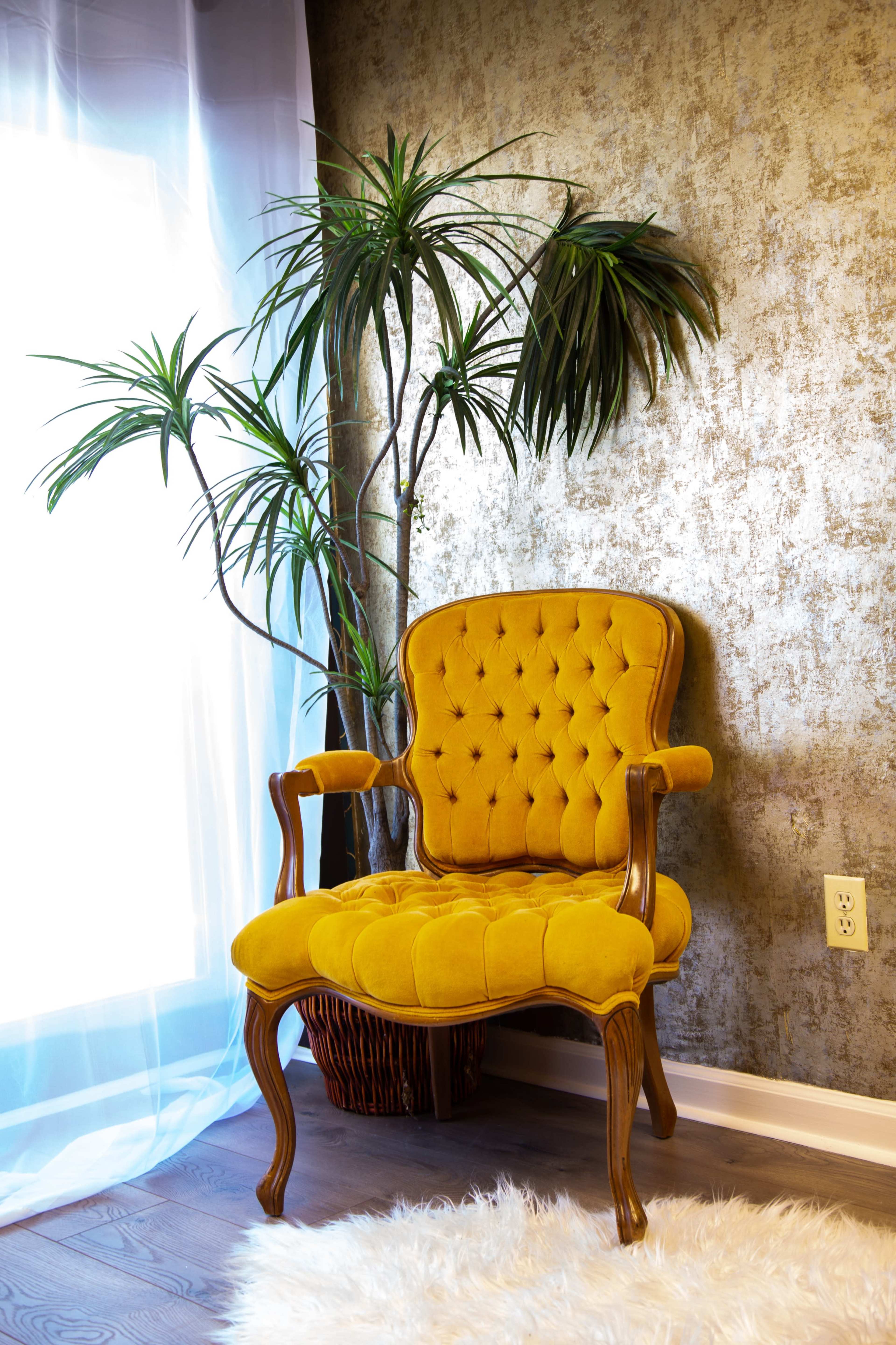 A yellow upholstered chair with tufted detailing sits near a window, accompanied by a leafy indoor plant against a textured wall.