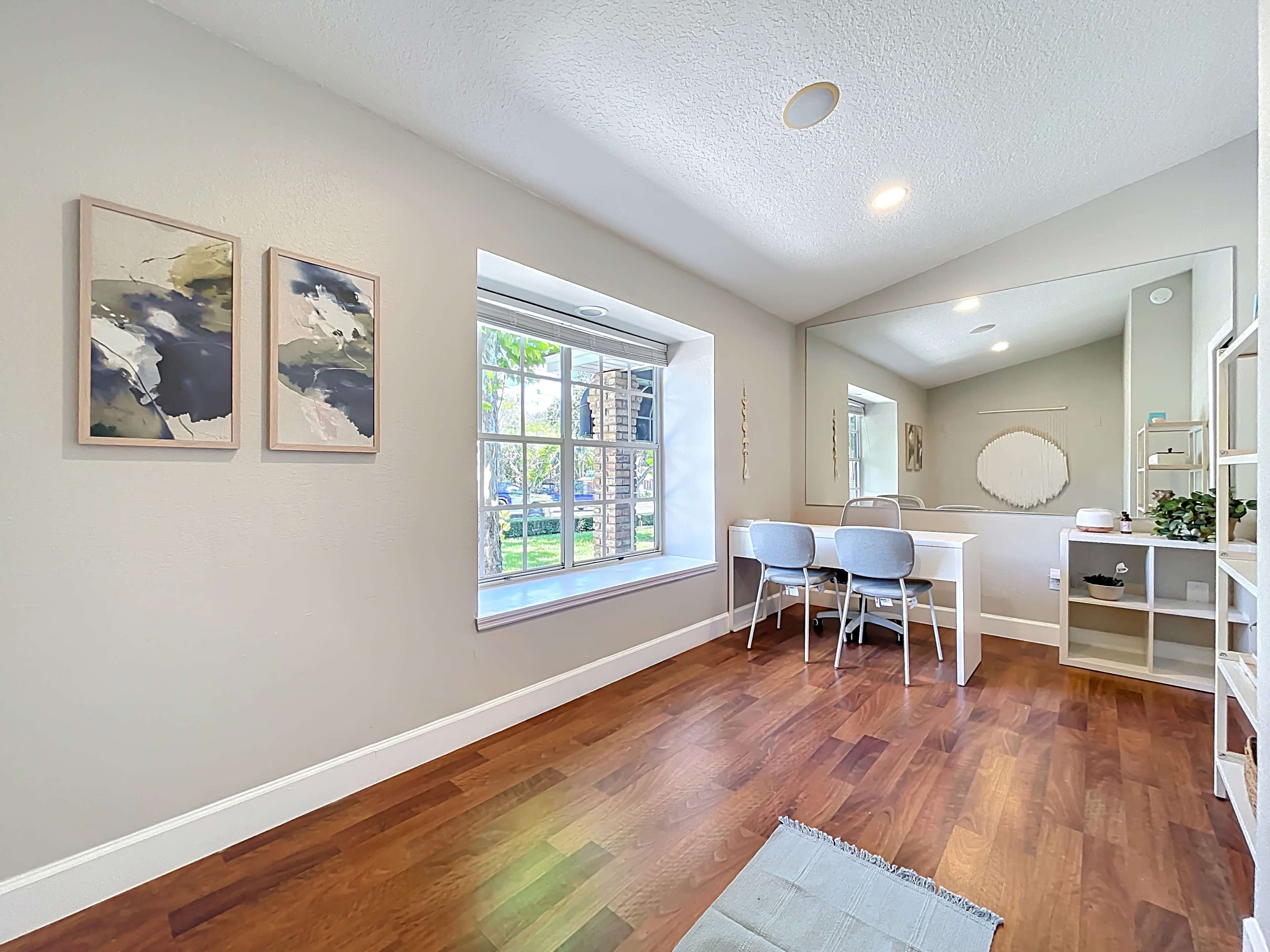 The image shows a bright, minimalist room featuring a window with a view, a wall-mounted mirror, and a small desk with two chairs.