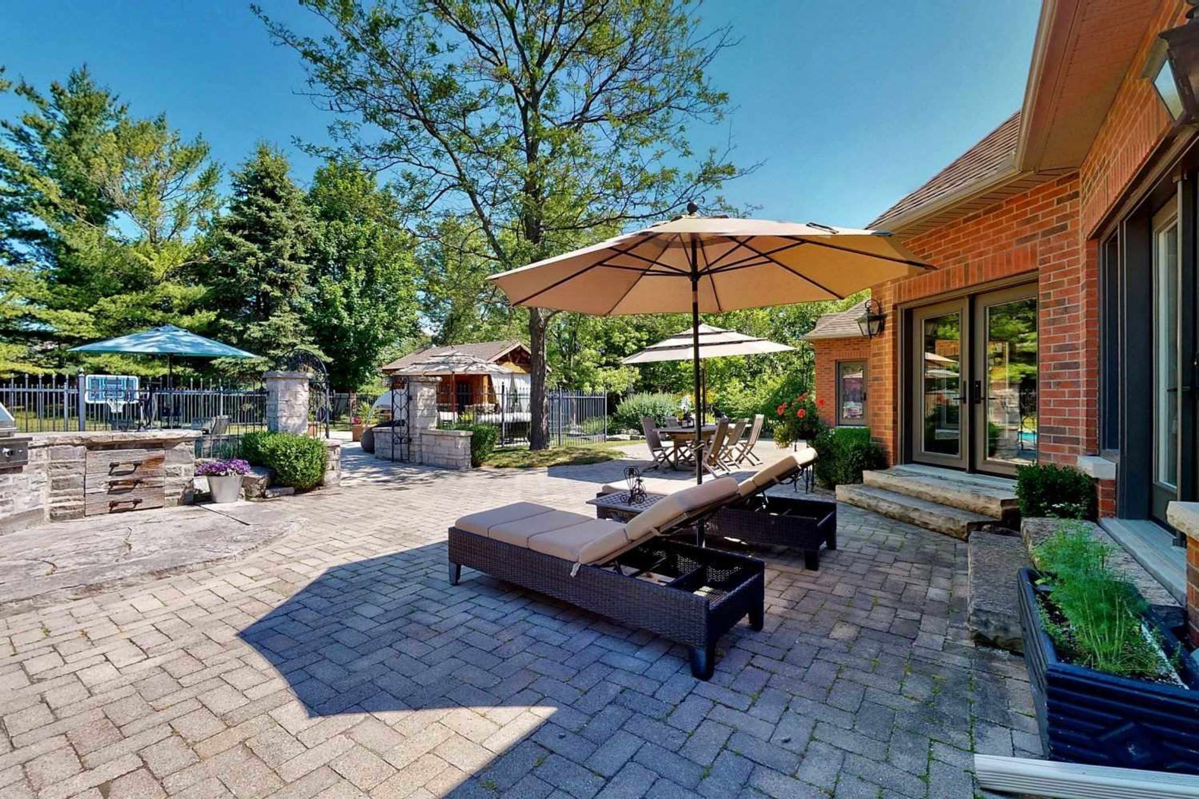 The image shows a stone patio featuring lounge chairs with umbrellas, surrounded by greenery and a barbecue area.