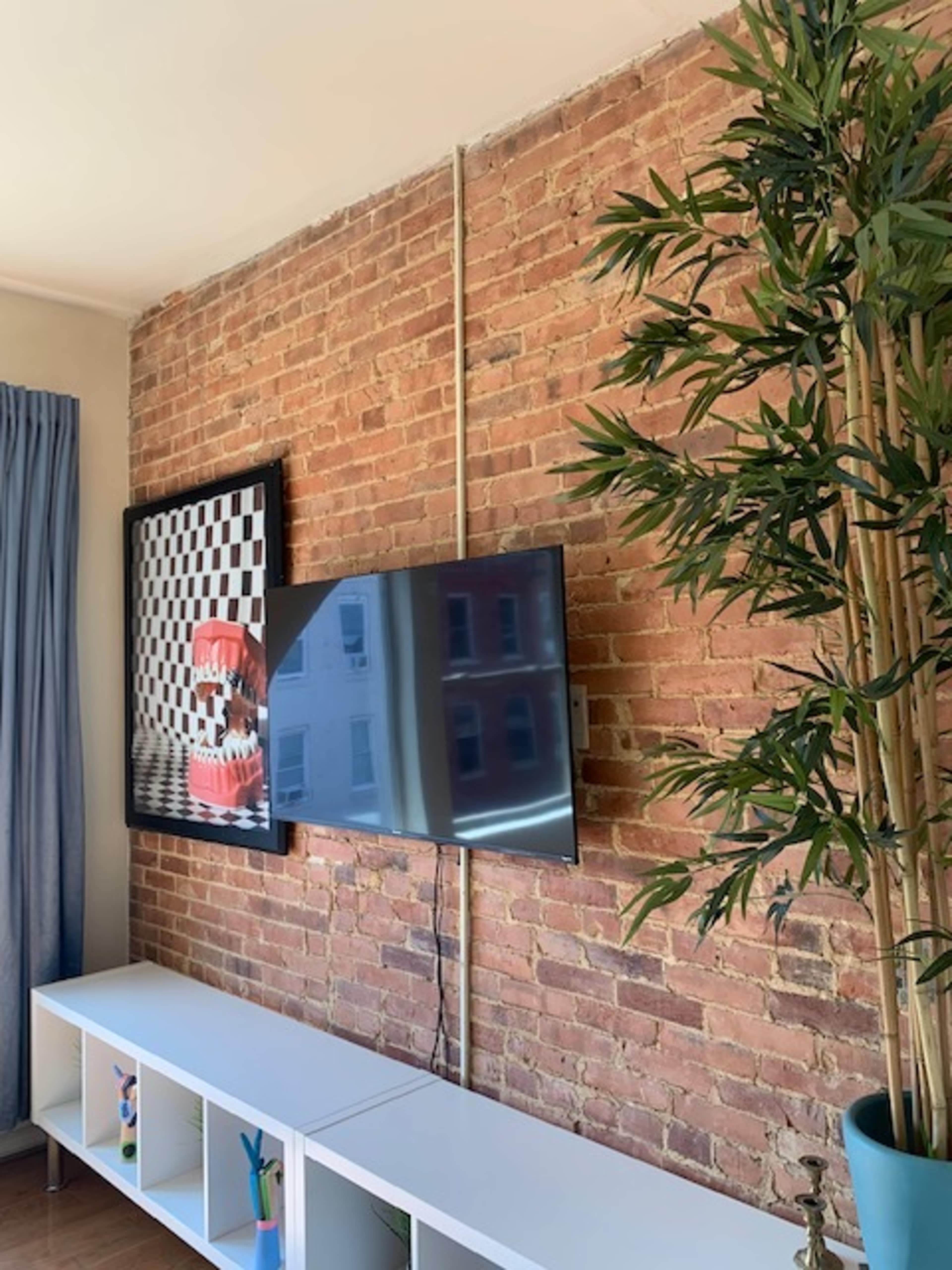 The image shows a modern living room featuring a wall with exposed brick, a large TV mounted on it, and a decorative potted plant beside a minimalist white shelf.