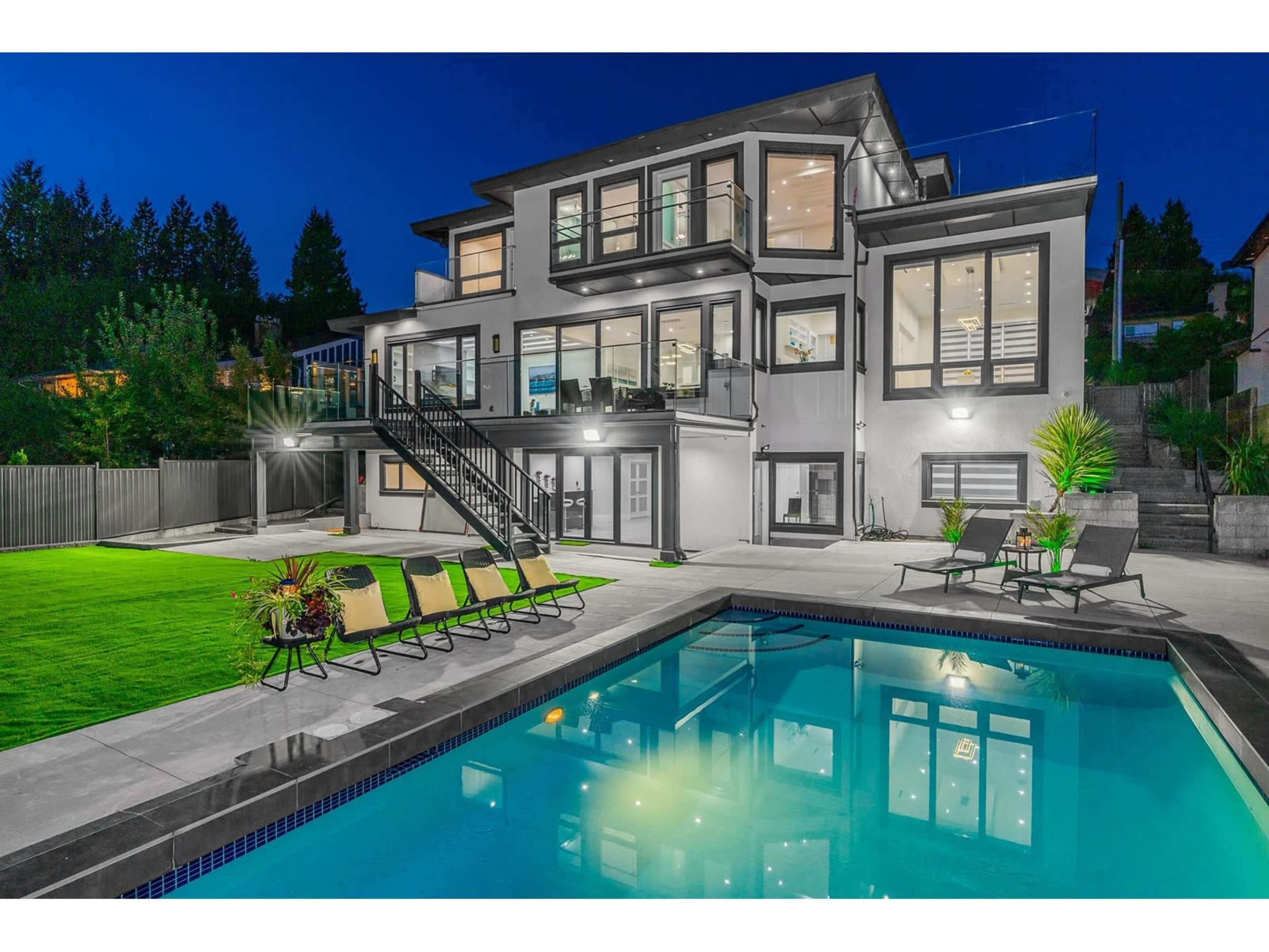 A modern two-story house with large windows overlooks a pool and landscaped yard, illuminated by exterior lights at night.