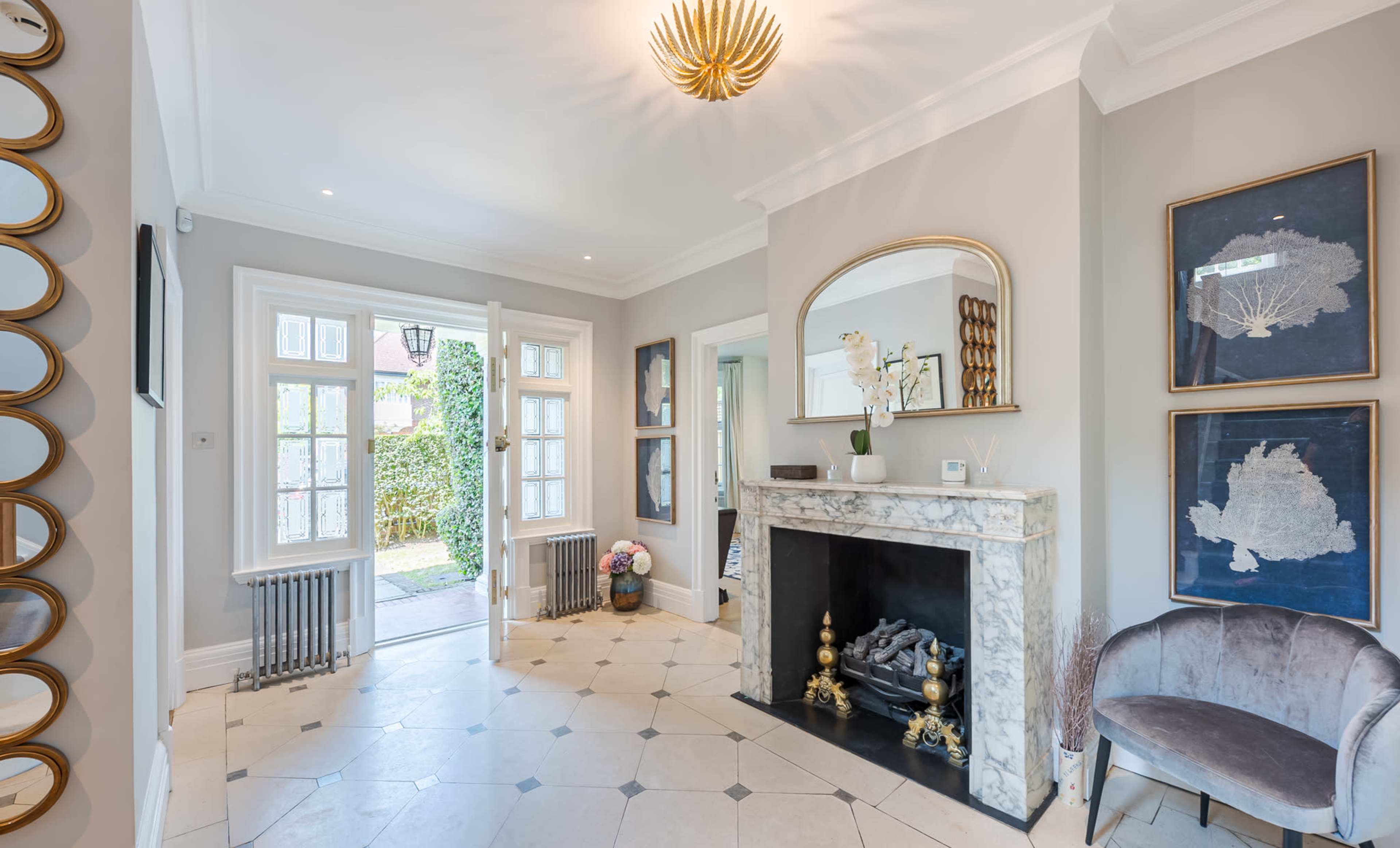 A well-lit entryway featuring a marble fireplace, a round mirror, and decorative artwork on the walls, with doors opening to a garden outside.
