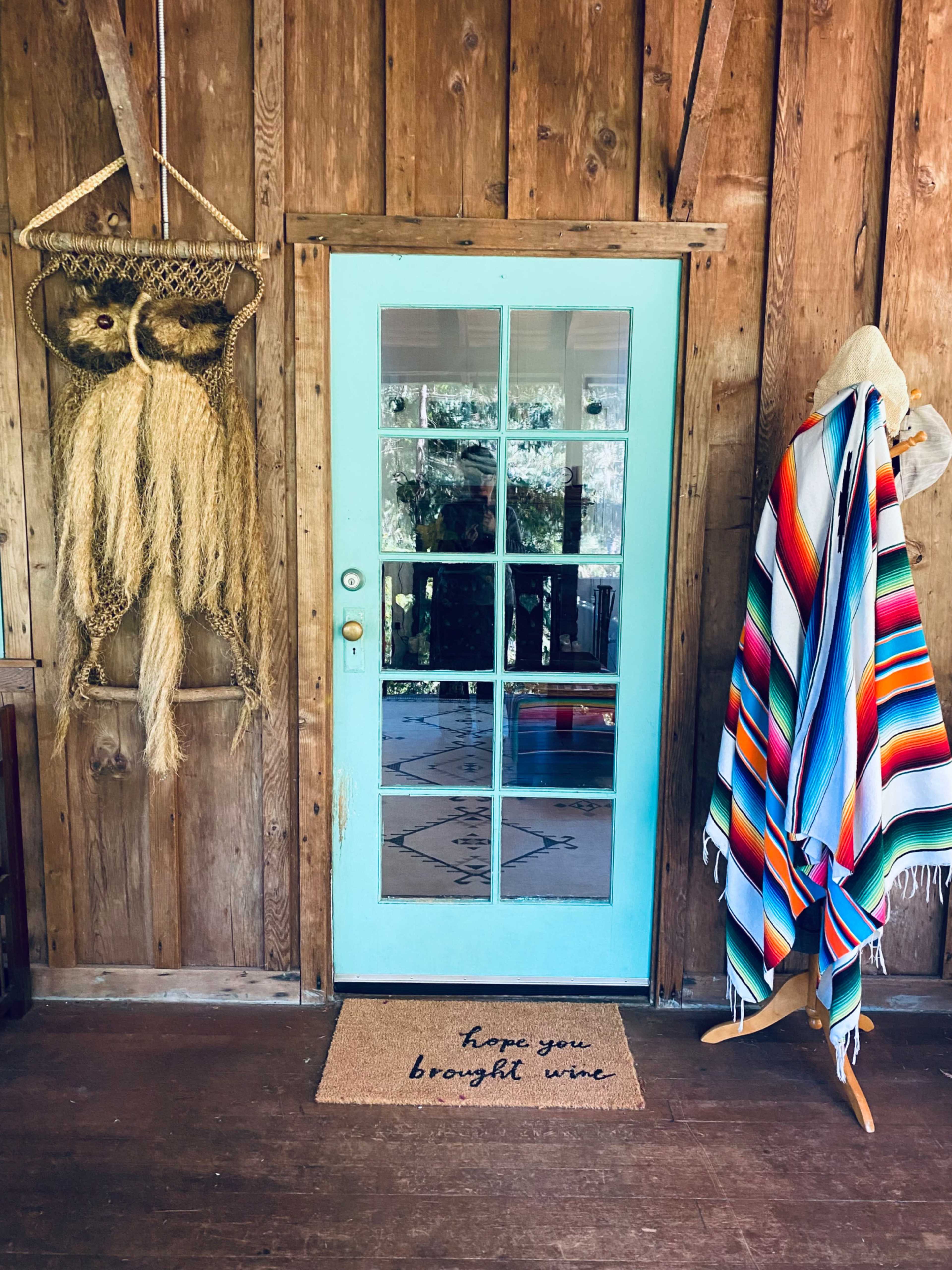 The image shows a turquoise door with glass panels, surrounded by wooden walls, a woven wall hanging, and a colorful striped blanket draped on a hook.