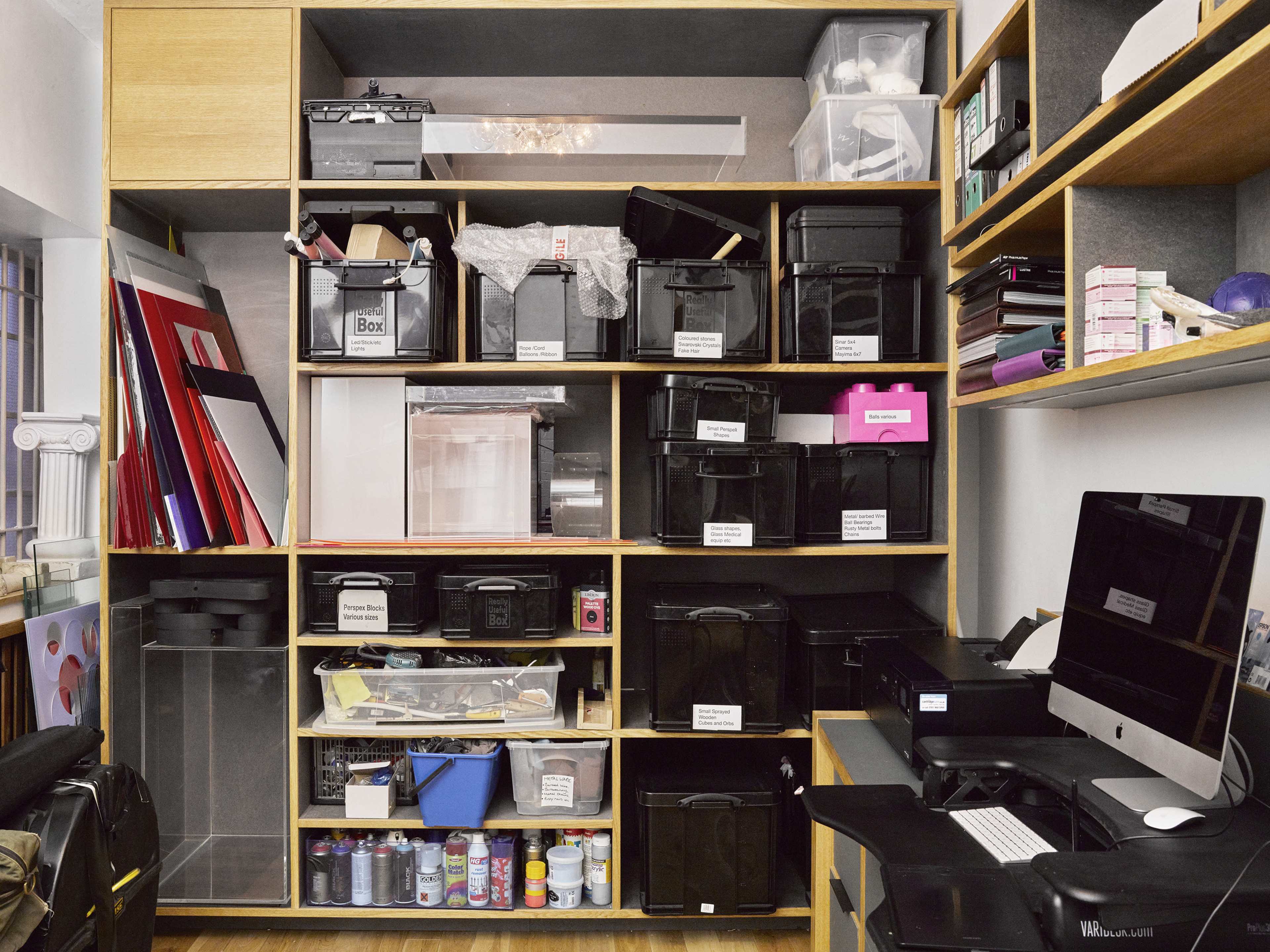 The image shows a neatly organized shelving unit filled with labeled storage bins, documents, and office supplies, alongside a computer setup on a black desk.
