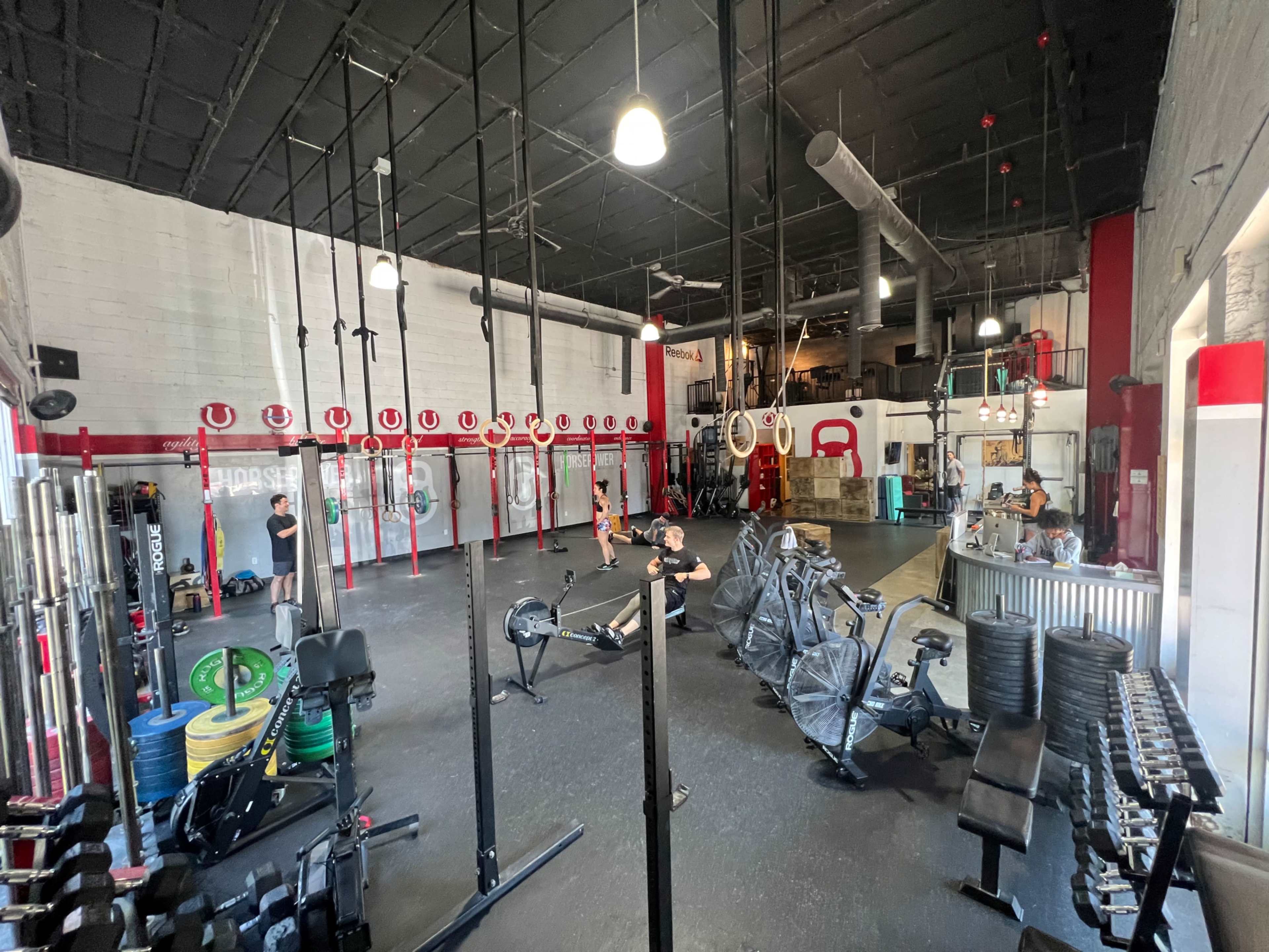 A spacious gym with various fitness equipment, including weights, rowing machines, and gymnastic rings, with several people exercising.