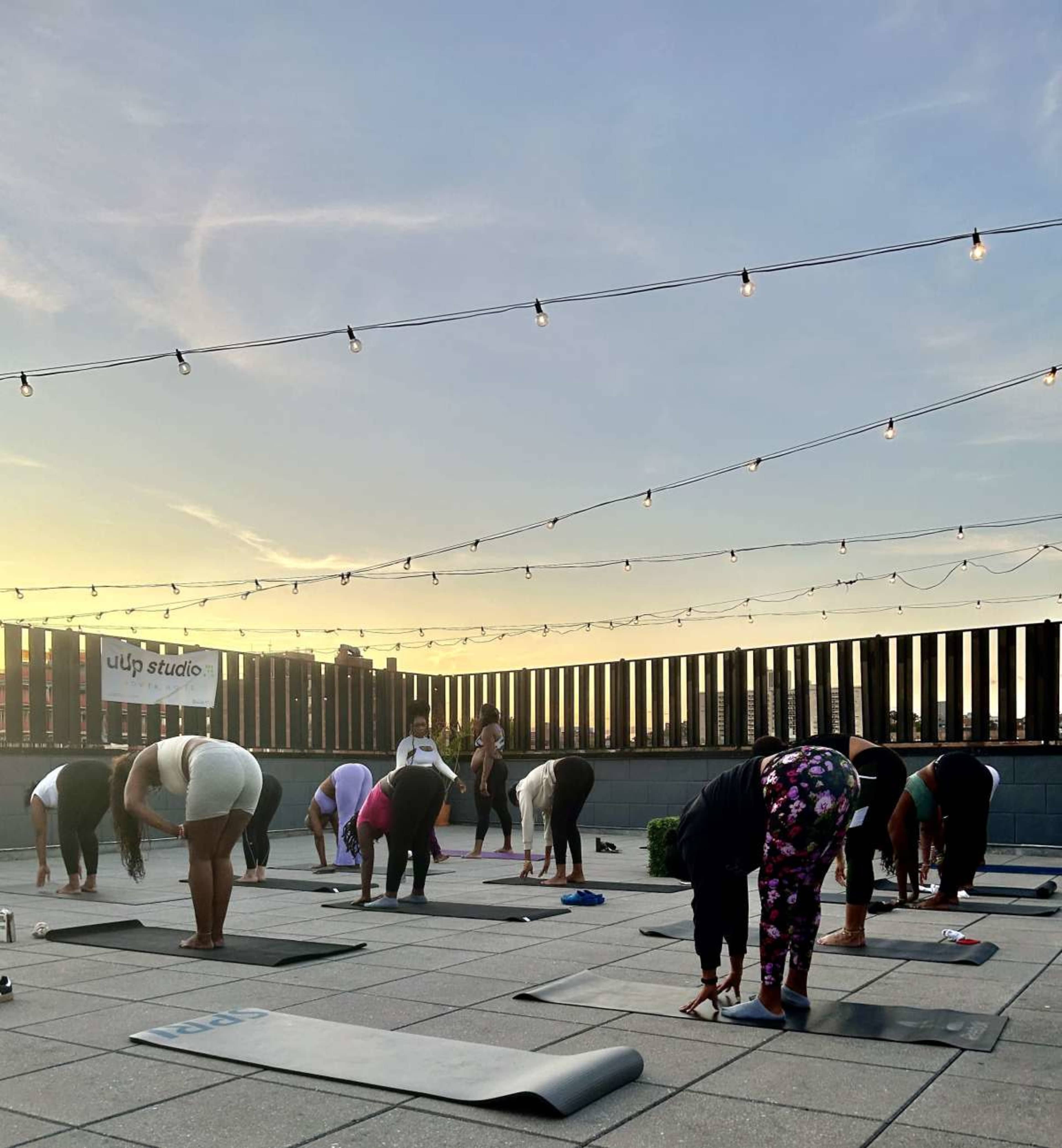 A group of people is practicing yoga on mats on a rooftop during sunset, with string lights overhead.