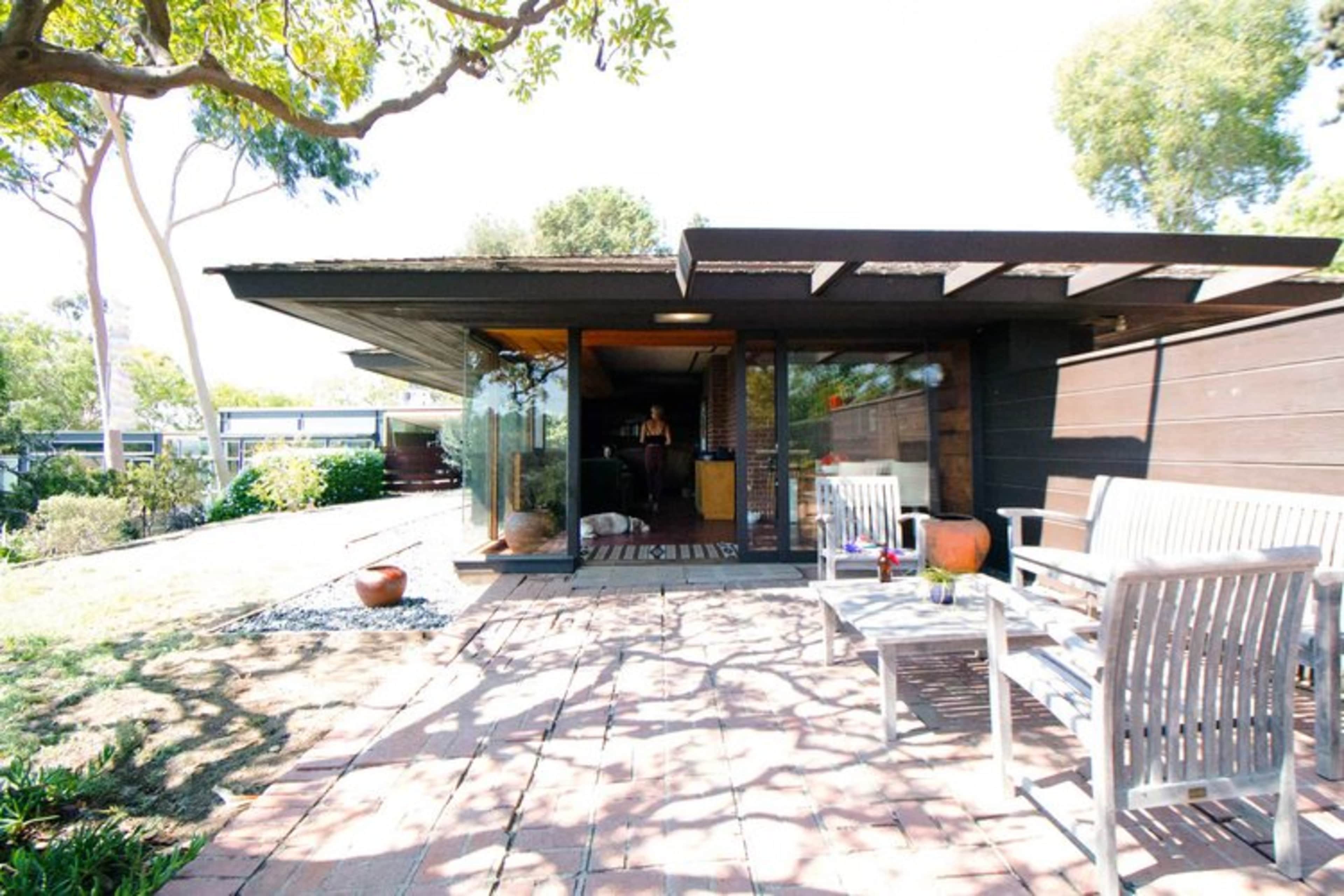 A modern house with a flat roof and large glass doors opens to a patio surrounded by trees and a well-maintained garden.
