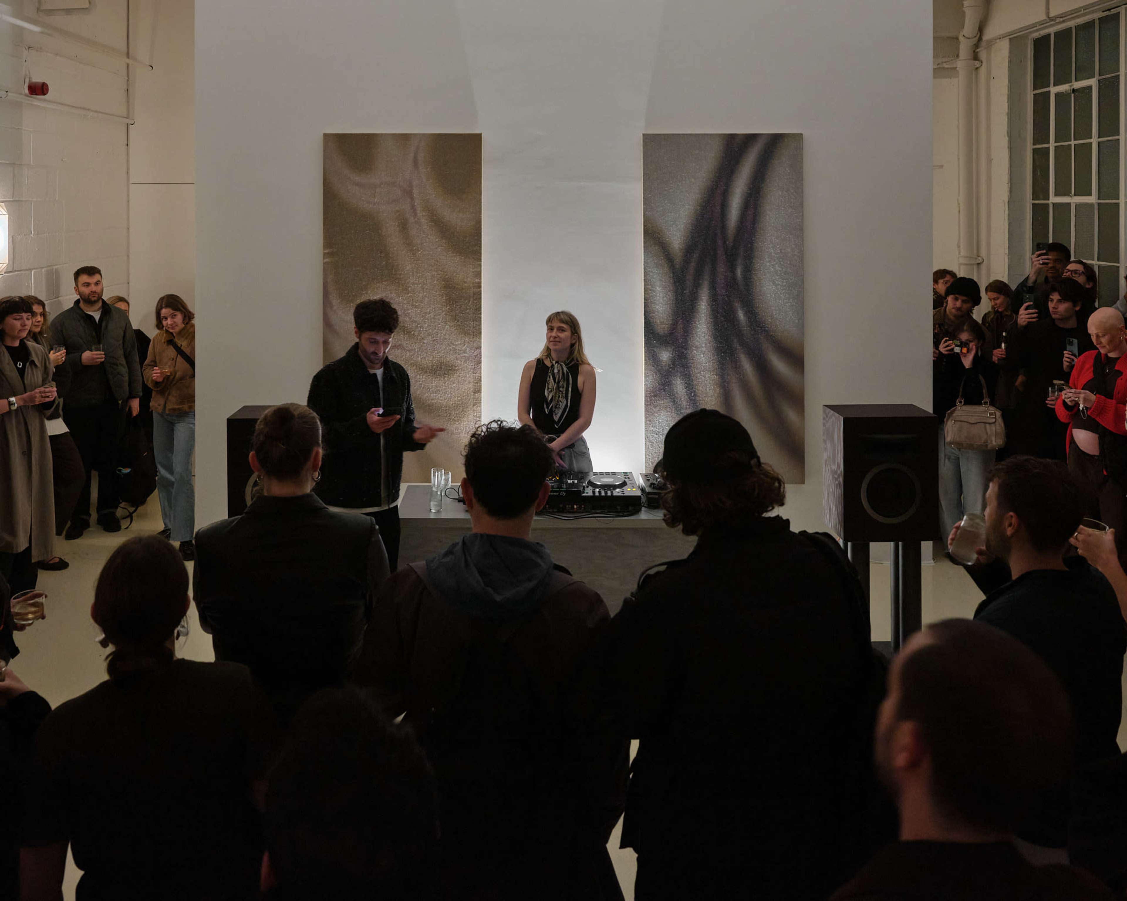 A crowd gathers around a DJ set in an art gallery, where a female DJ performs while a male speaker addresses the audience.