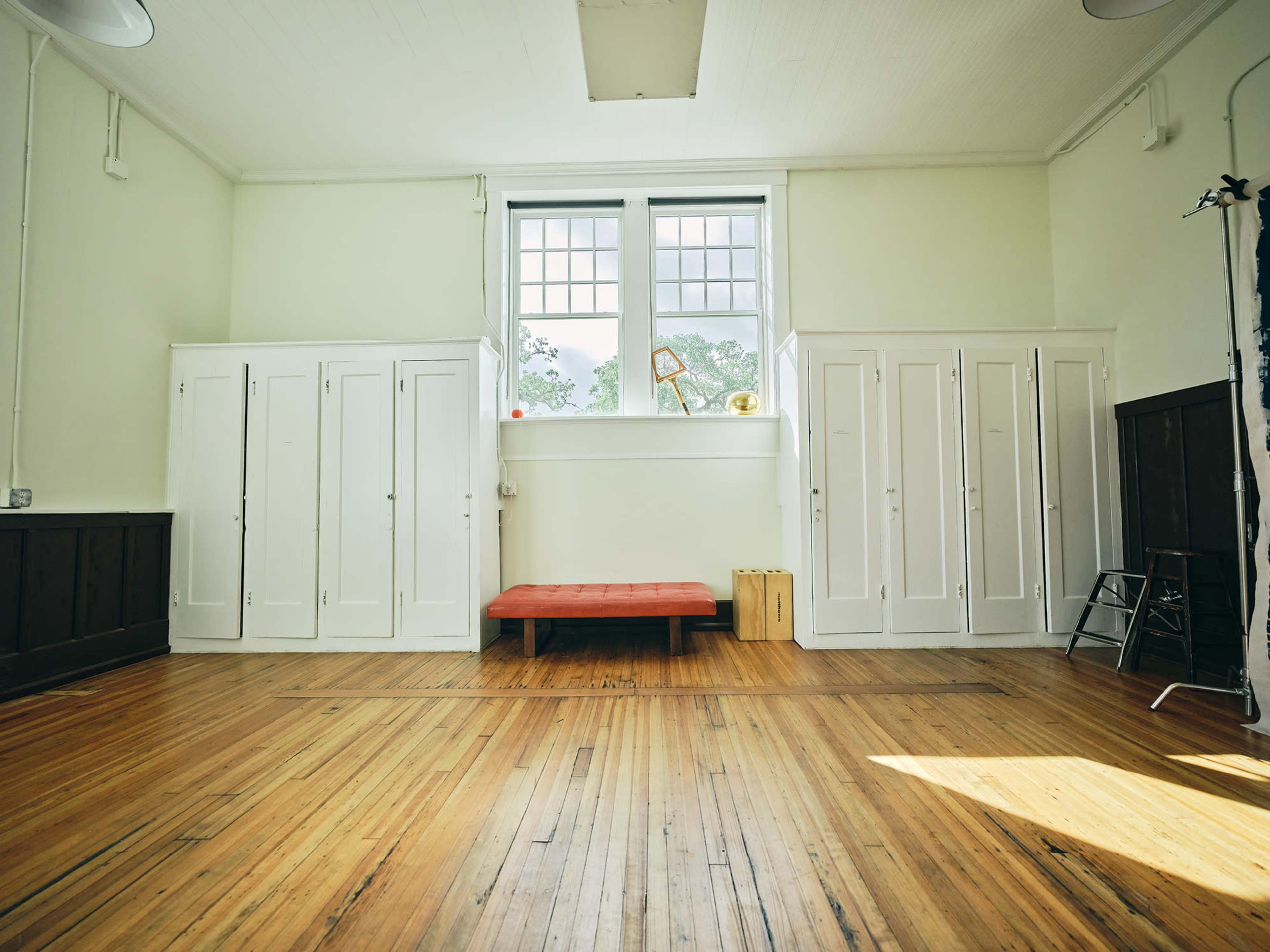 The image shows a spacious room with wooden floors, two sets of white storage cabinets on either side, and a red bench in the center near large windows.