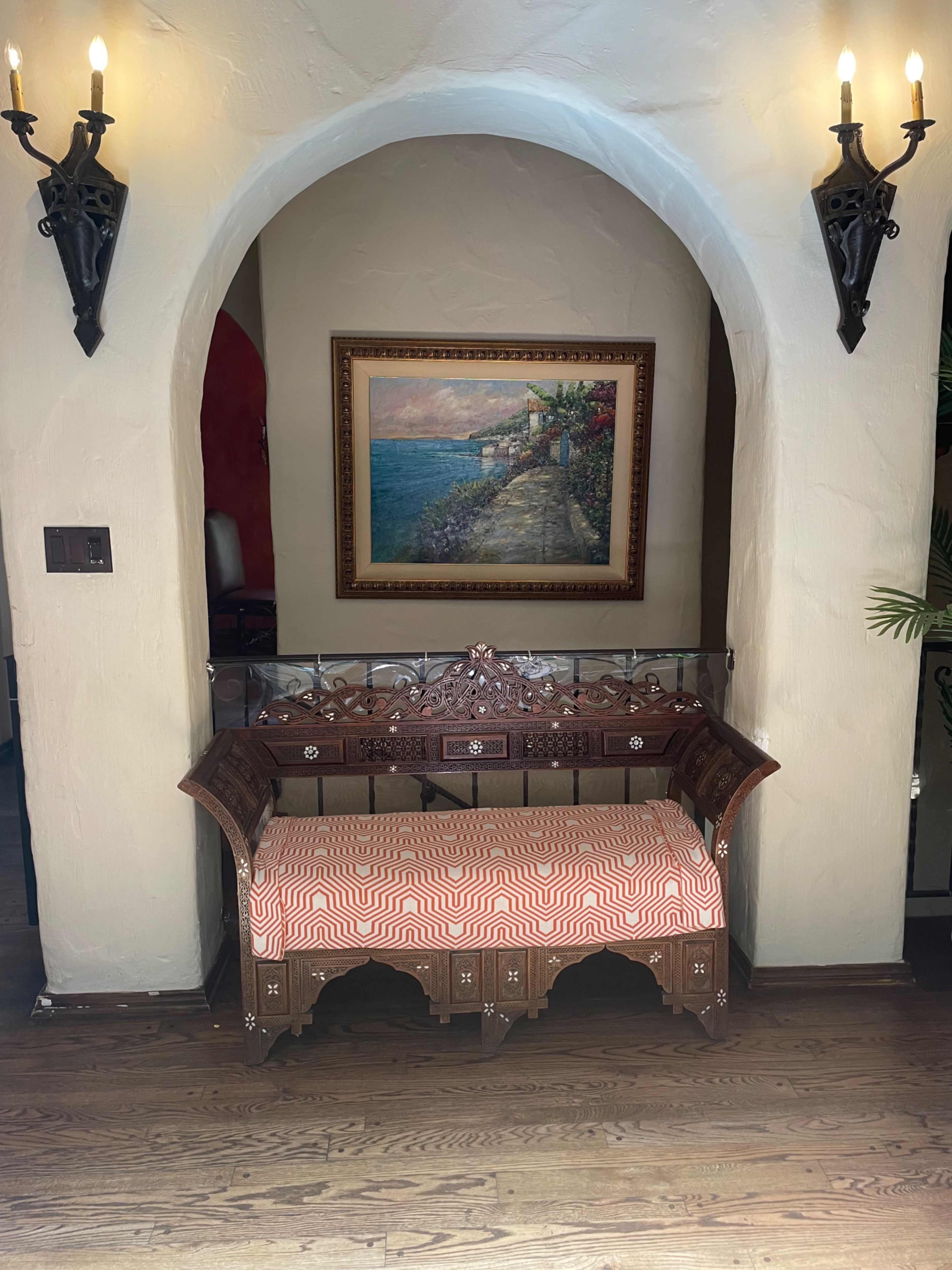 A decorative wooden bench with an upholstered seat, positioned under an archway with wall sconces and a framed landscape painting in the background.