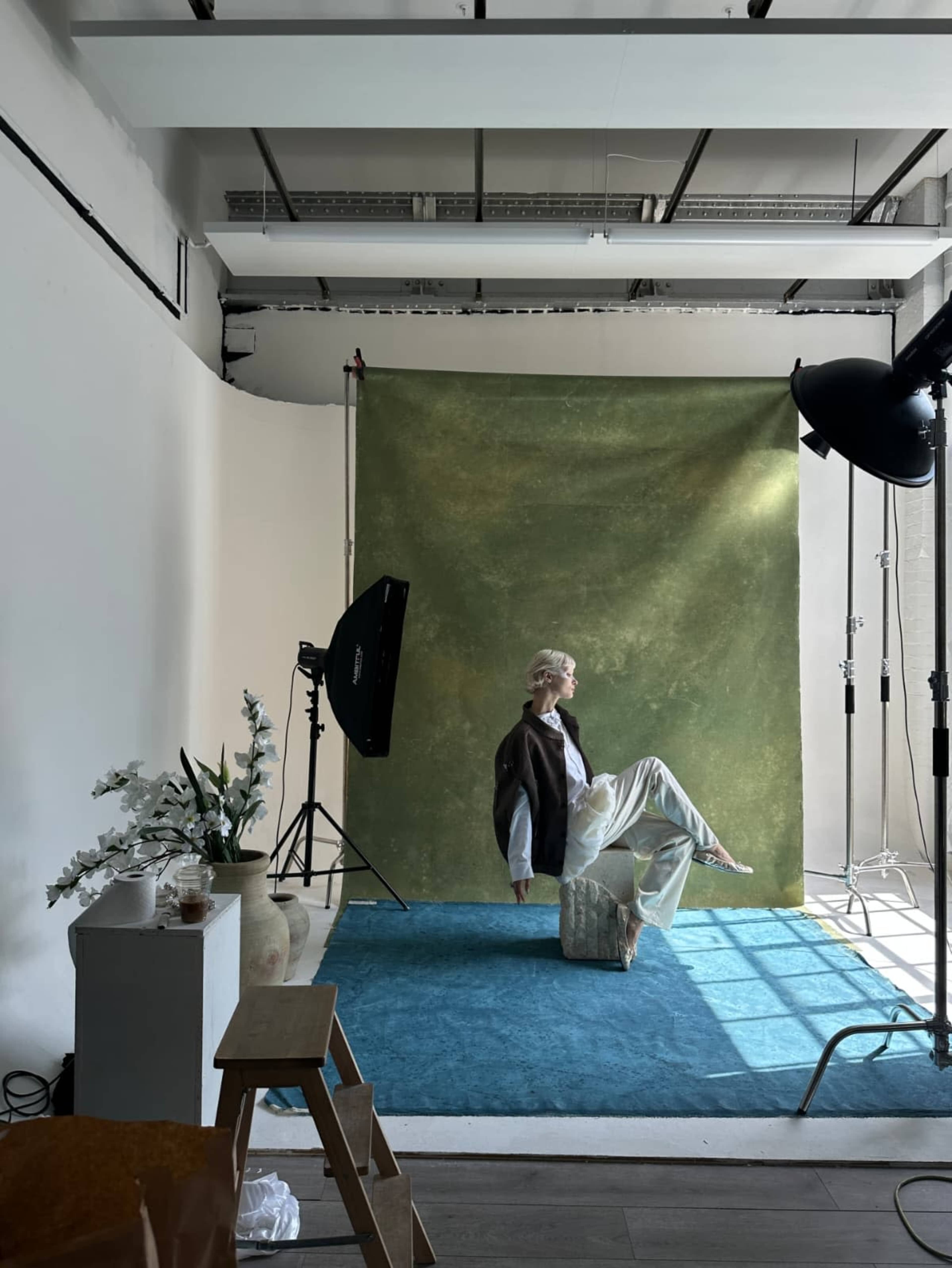 A model poses on a stone in front of a green backdrop in a photography studio, with lighting equipment and plants in the scene.