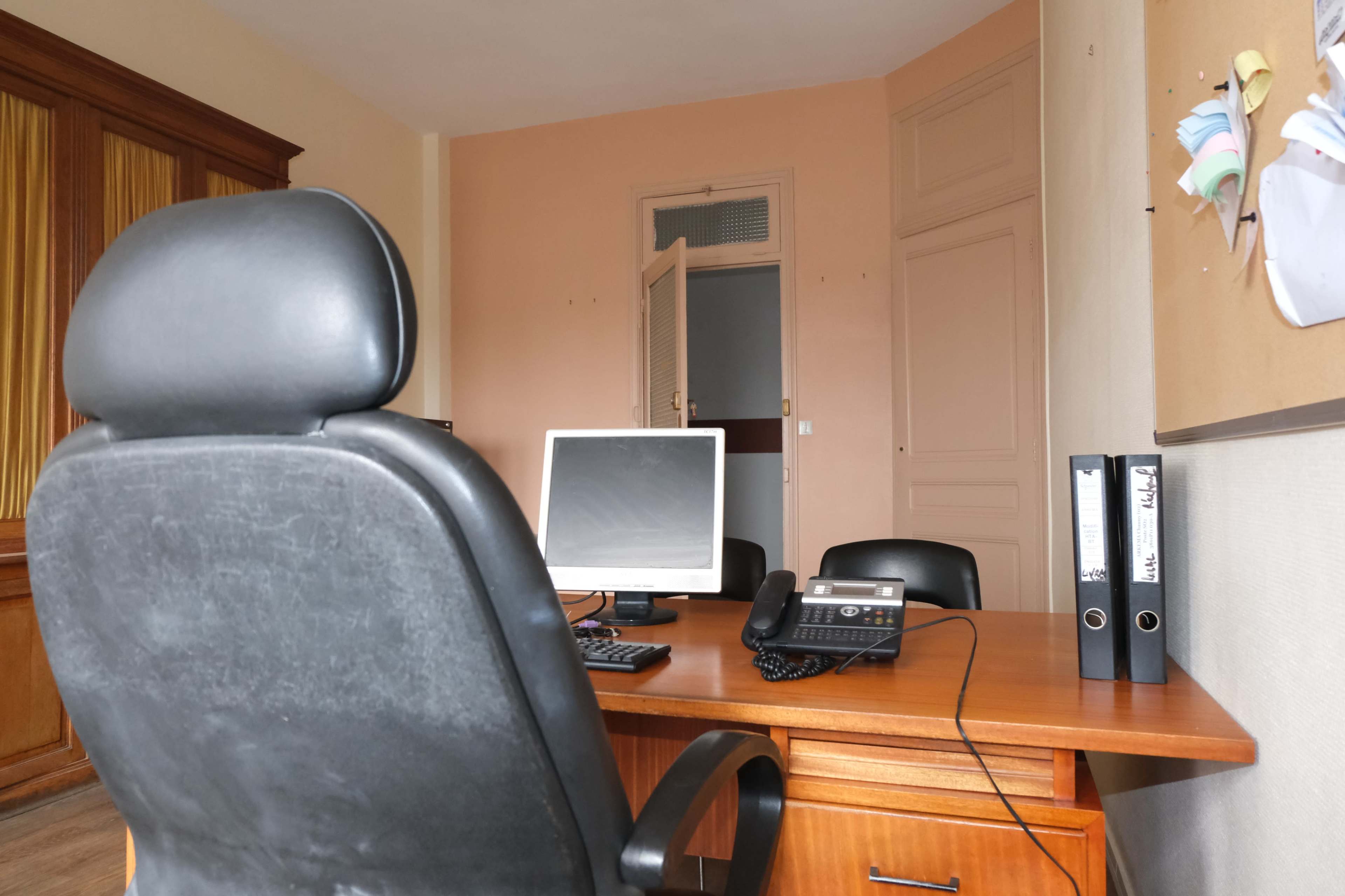 An office space is shown with a wooden desk, a computer monitor, a telephone, and black chairs, with a bulletin board on the wall.