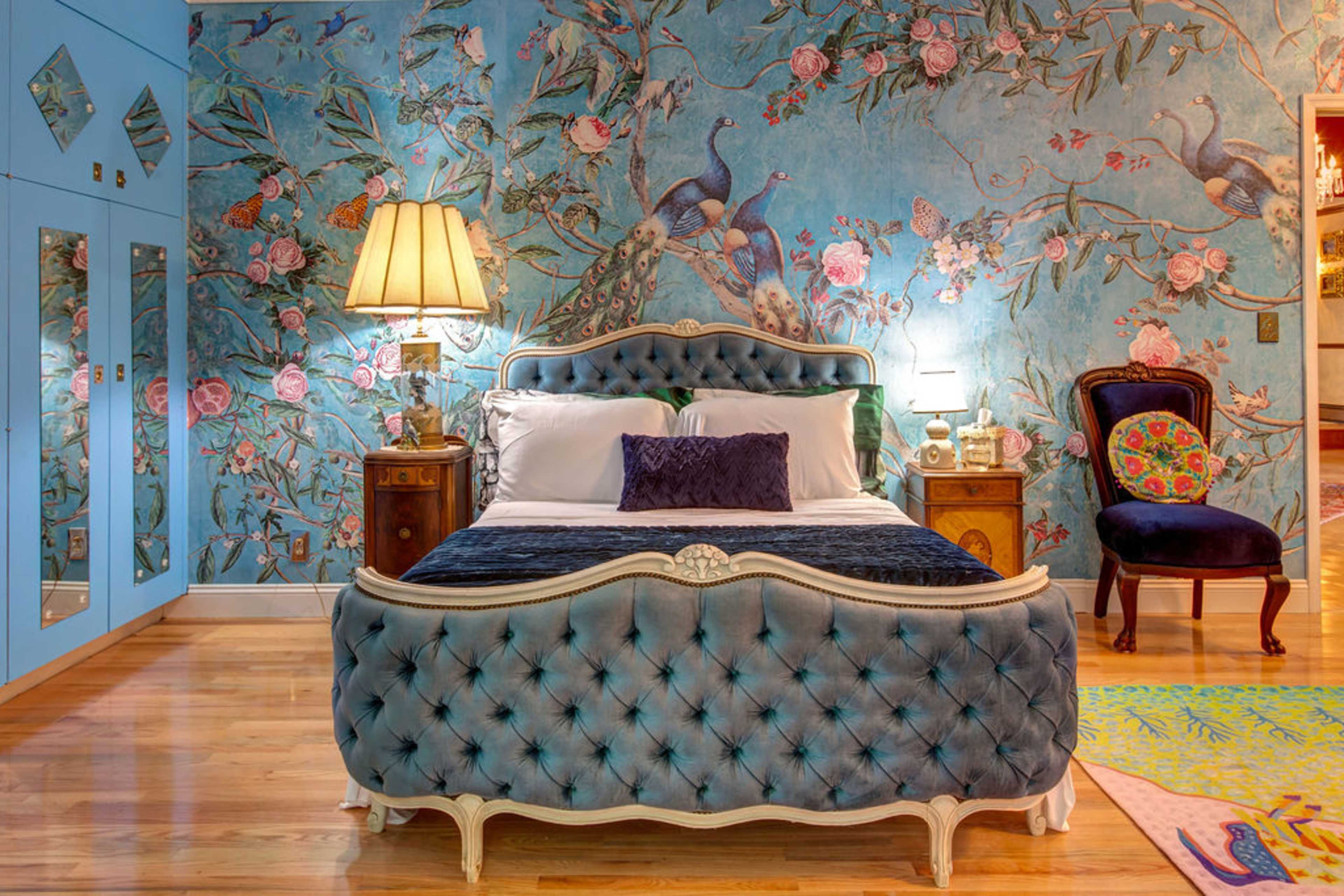 The image shows a bedroom featuring a blue tufted bed with a decorative headboard, a floral wallpaper backdrop, and a combination of luxurious furniture elements.
