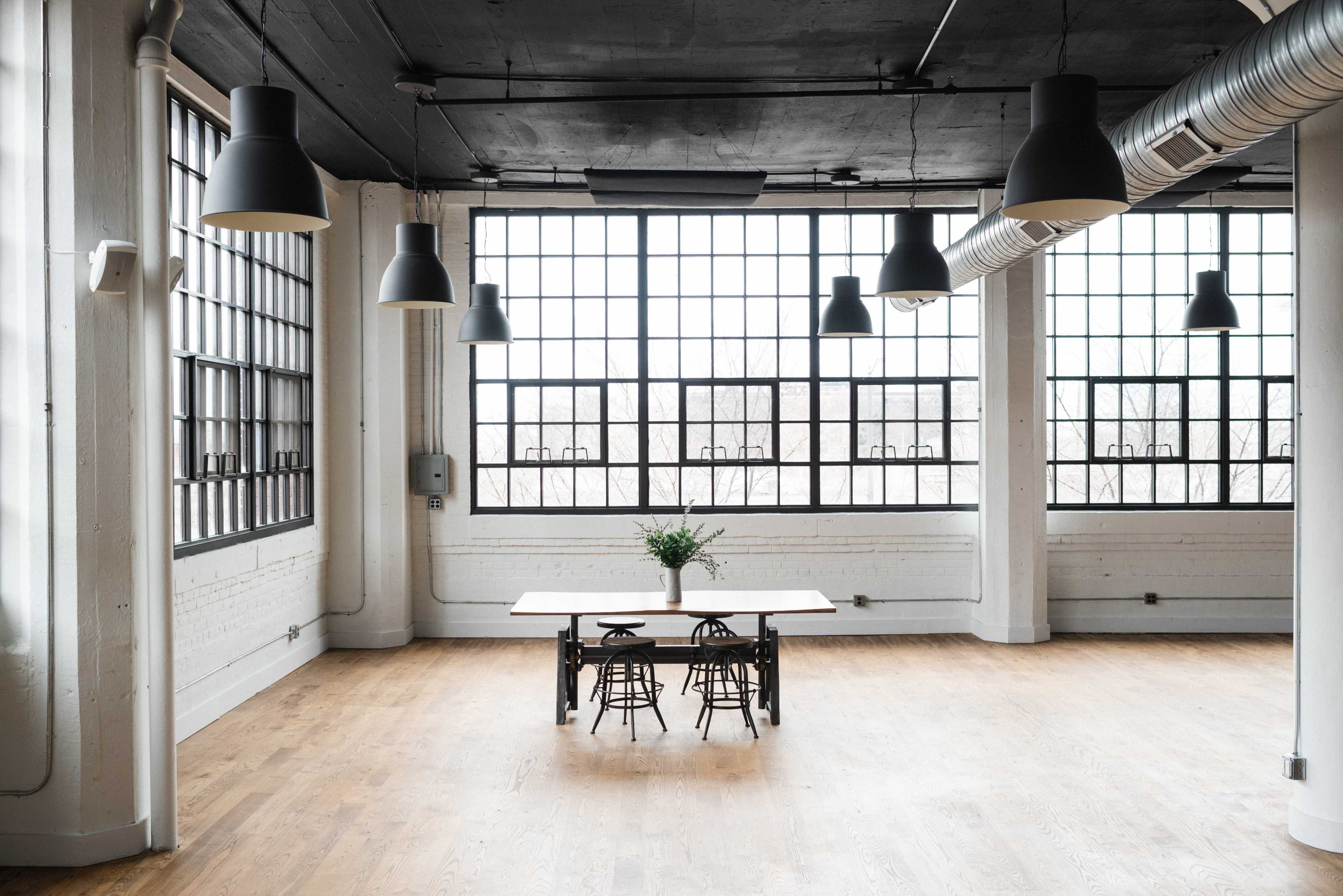 A spacious, bright room features a central wooden table surrounded by metal chairs, with large windows lining the walls.