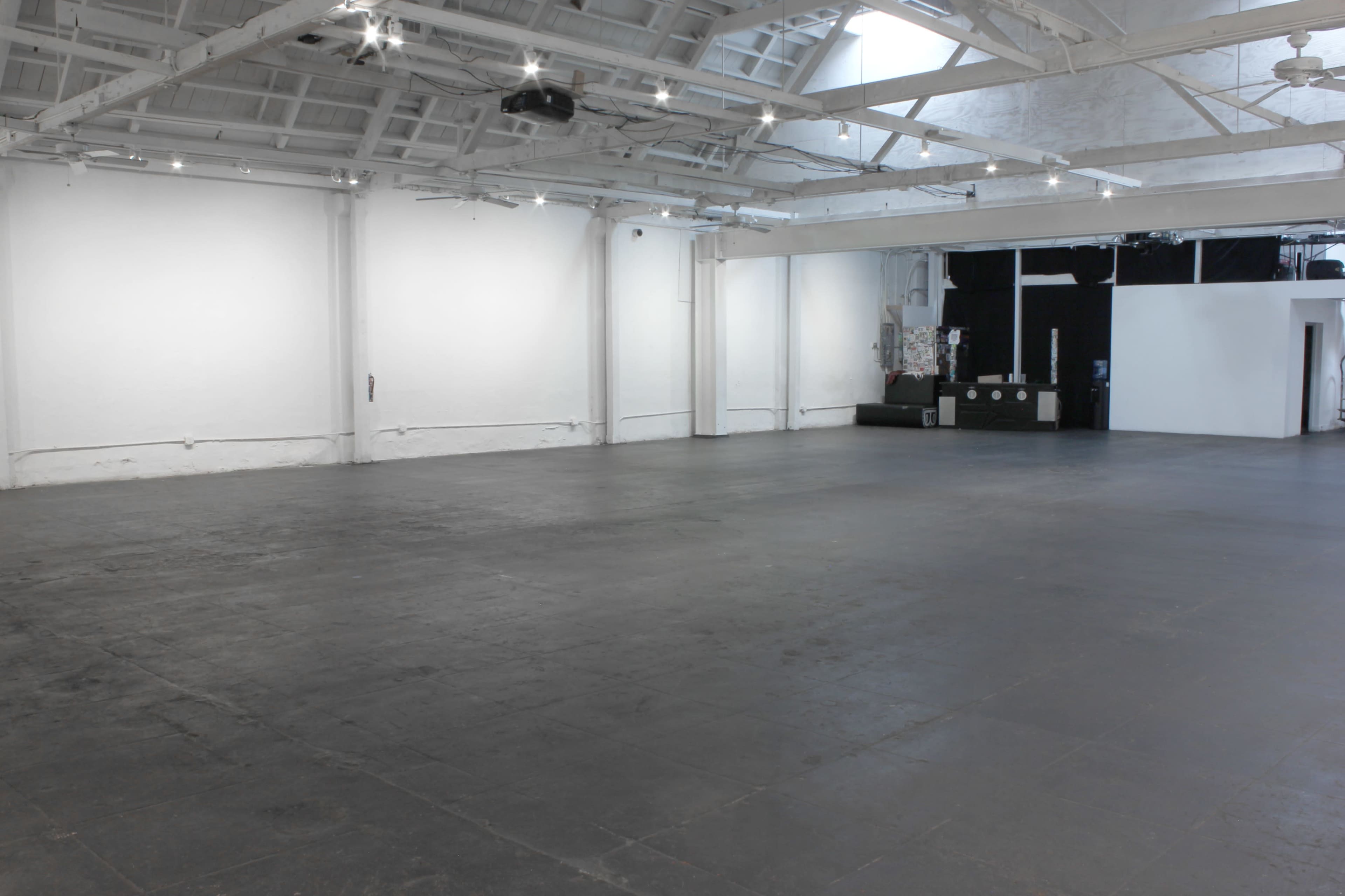 The image shows a spacious, empty room with white walls, a high ceiling, and a black floor.