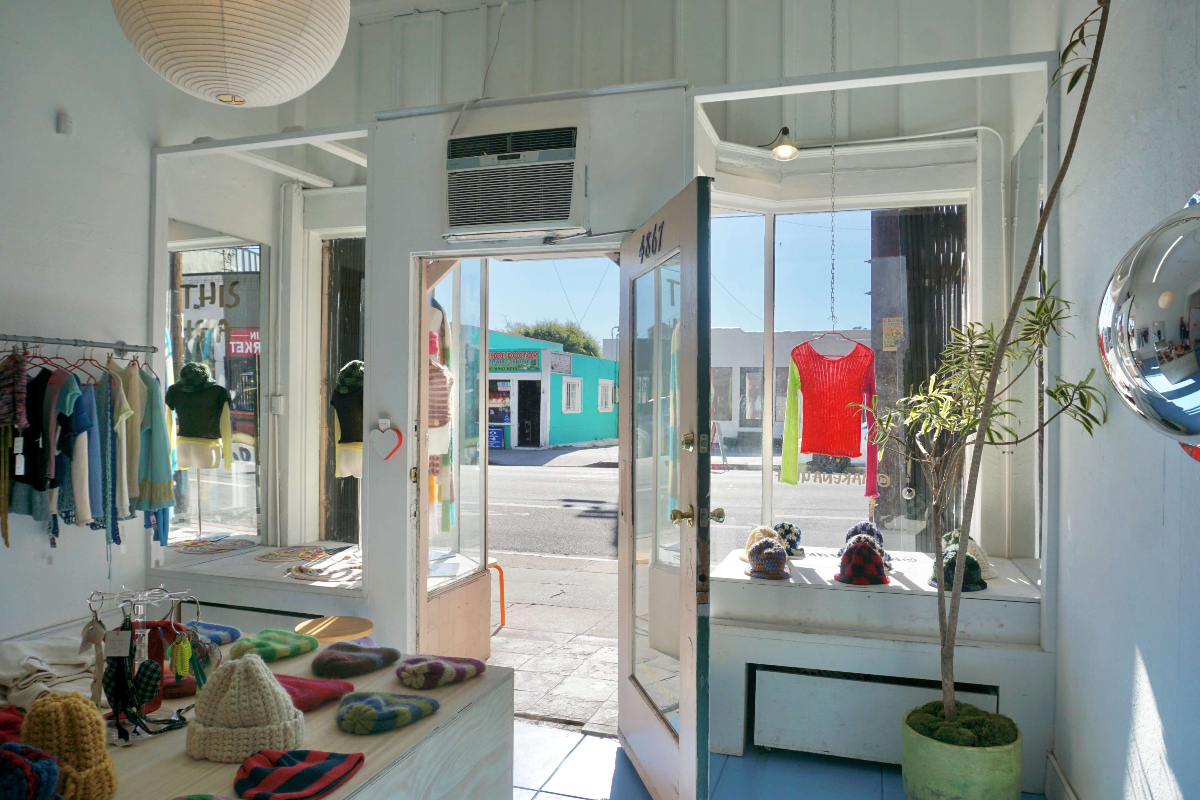The image shows the interior of a retail store with a doorway open to the street, displaying colorful knitwear and a small plant near the entrance.