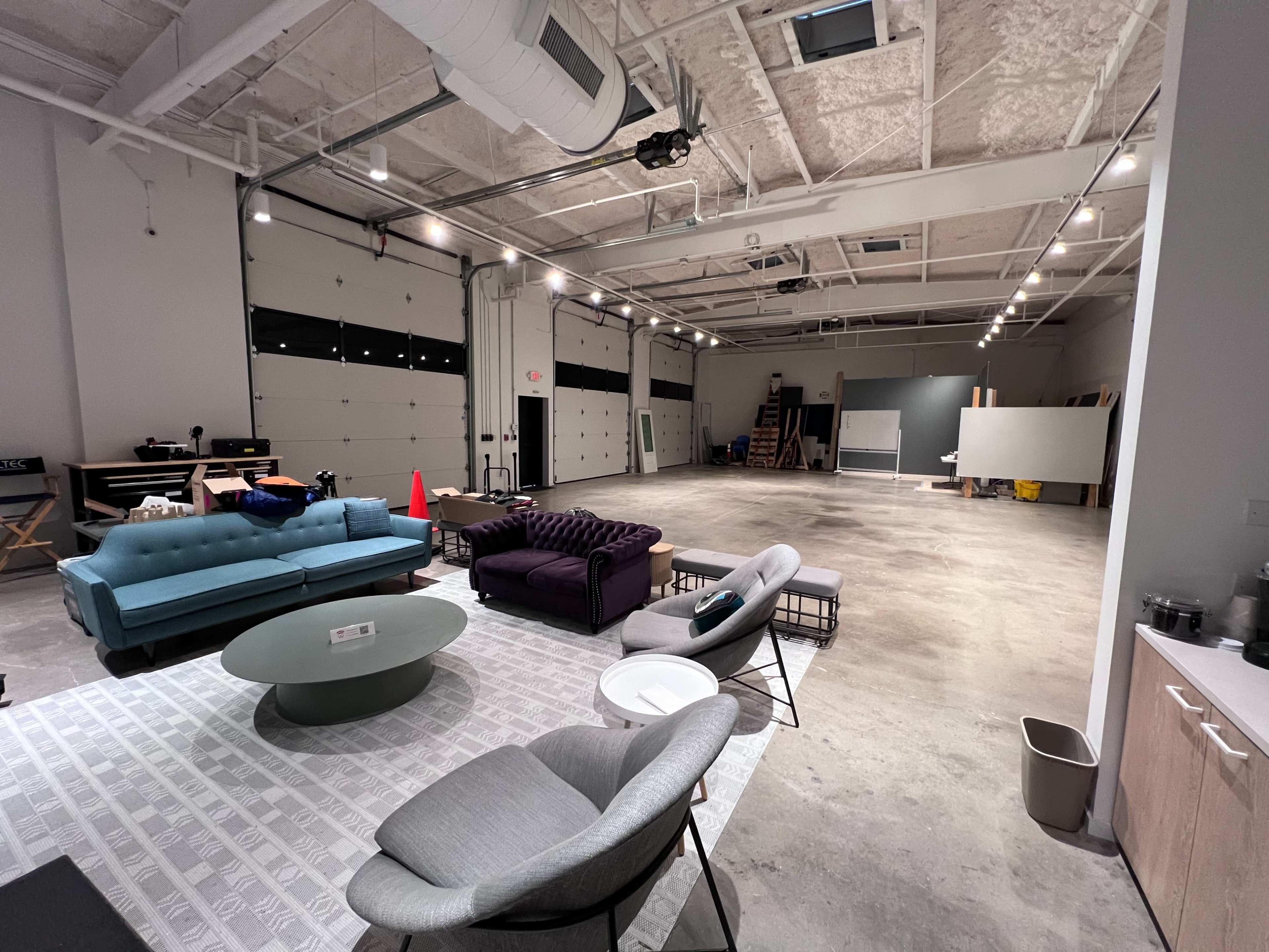 The image shows a spacious interior featuring a lounge area with various seating arrangements, including a blue couch, purple chairs, and a coffee table, alongside an open warehouse-style space with large garage doors.