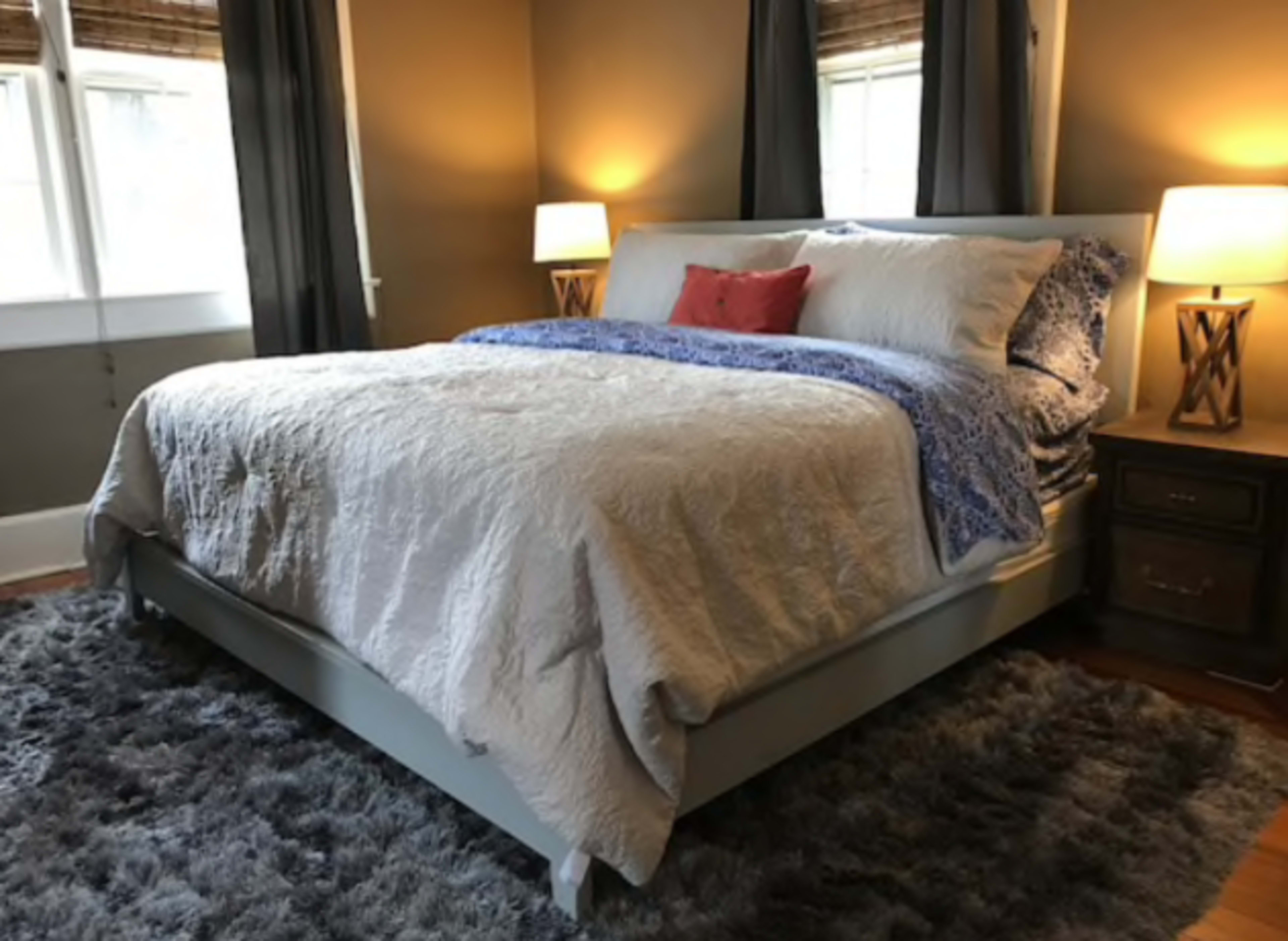 A well-made bed with a light-colored quilt and blue accent pillows, positioned in a warmly lit bedroom with two bedside lamps and dark curtains.