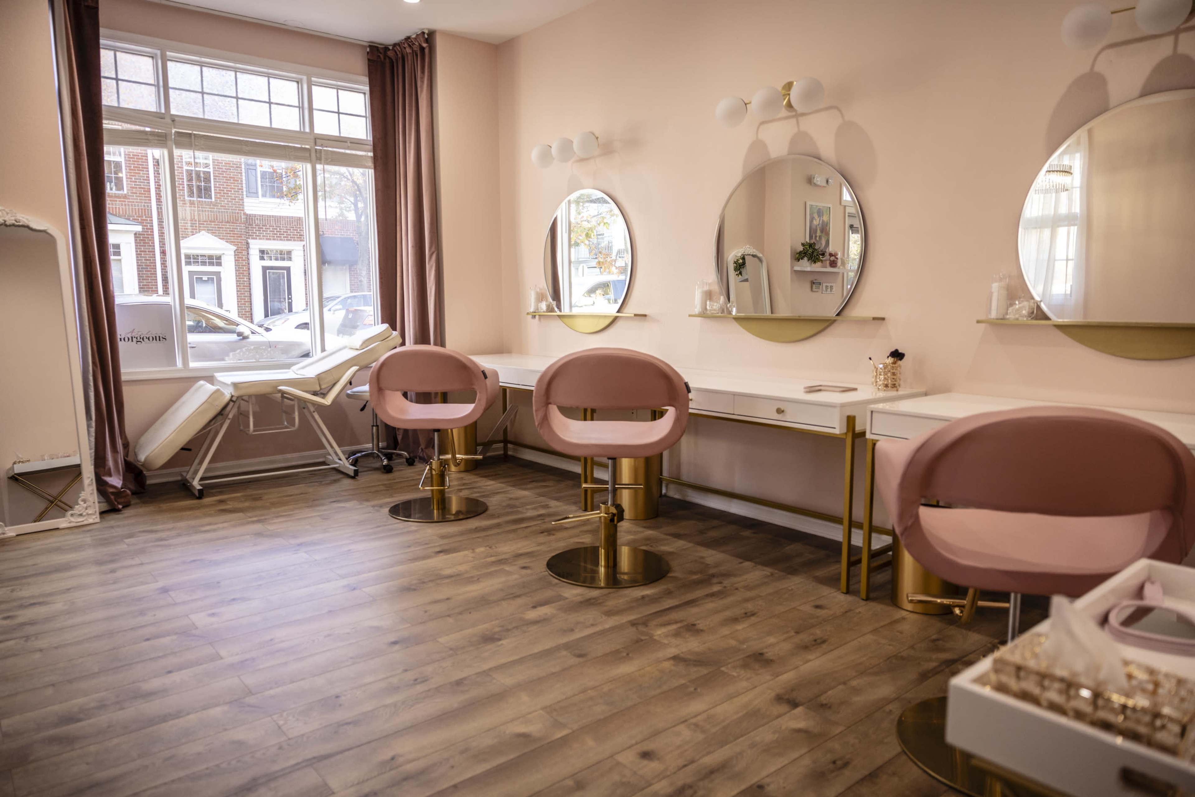 A beauty salon interior with three round mirrors and pink chairs arranged around white dressing tables under natural light from large windows.