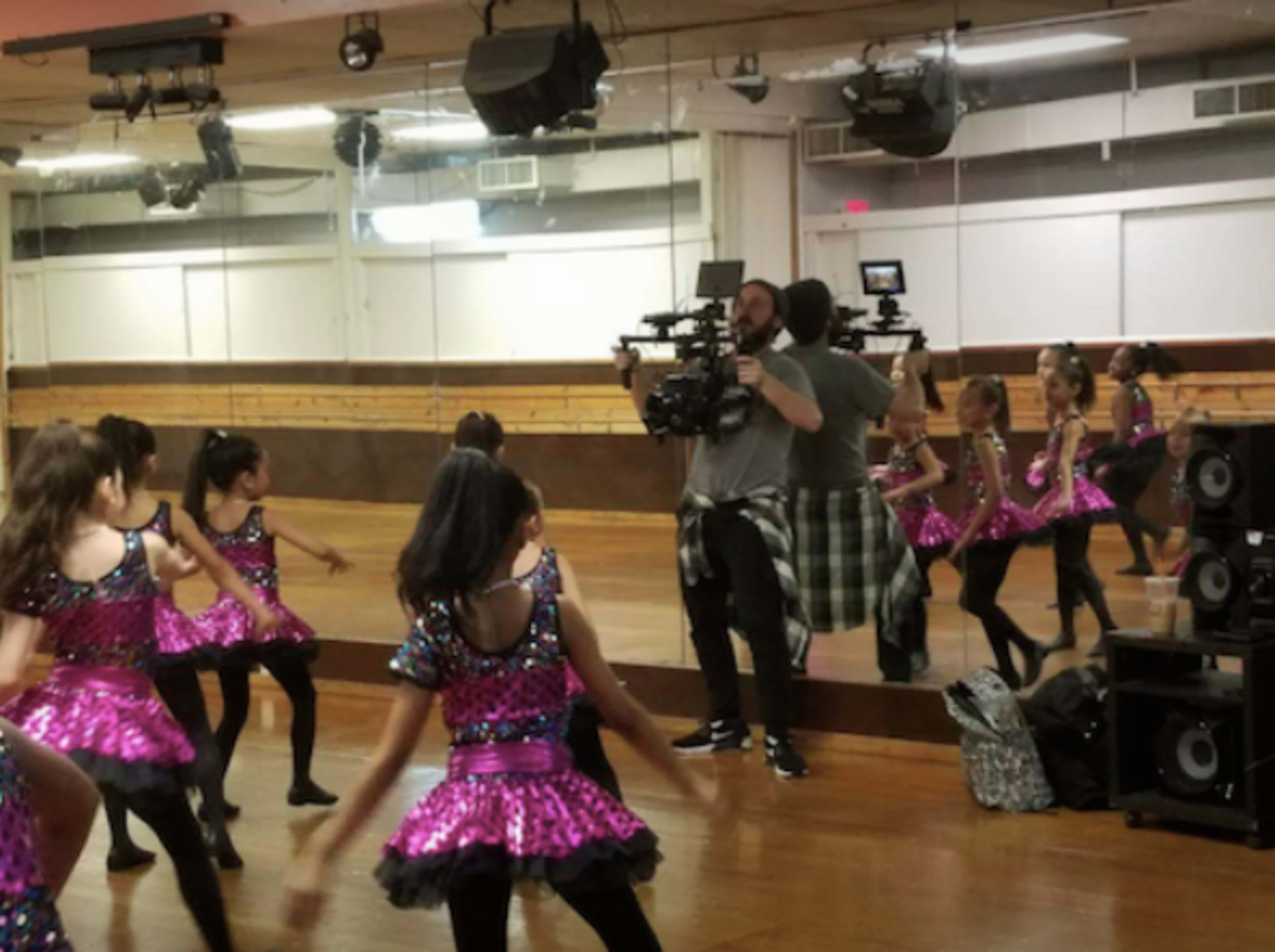 A group of children in sparkling pink costumes dances in a mirrored studio while a cameraman films them.