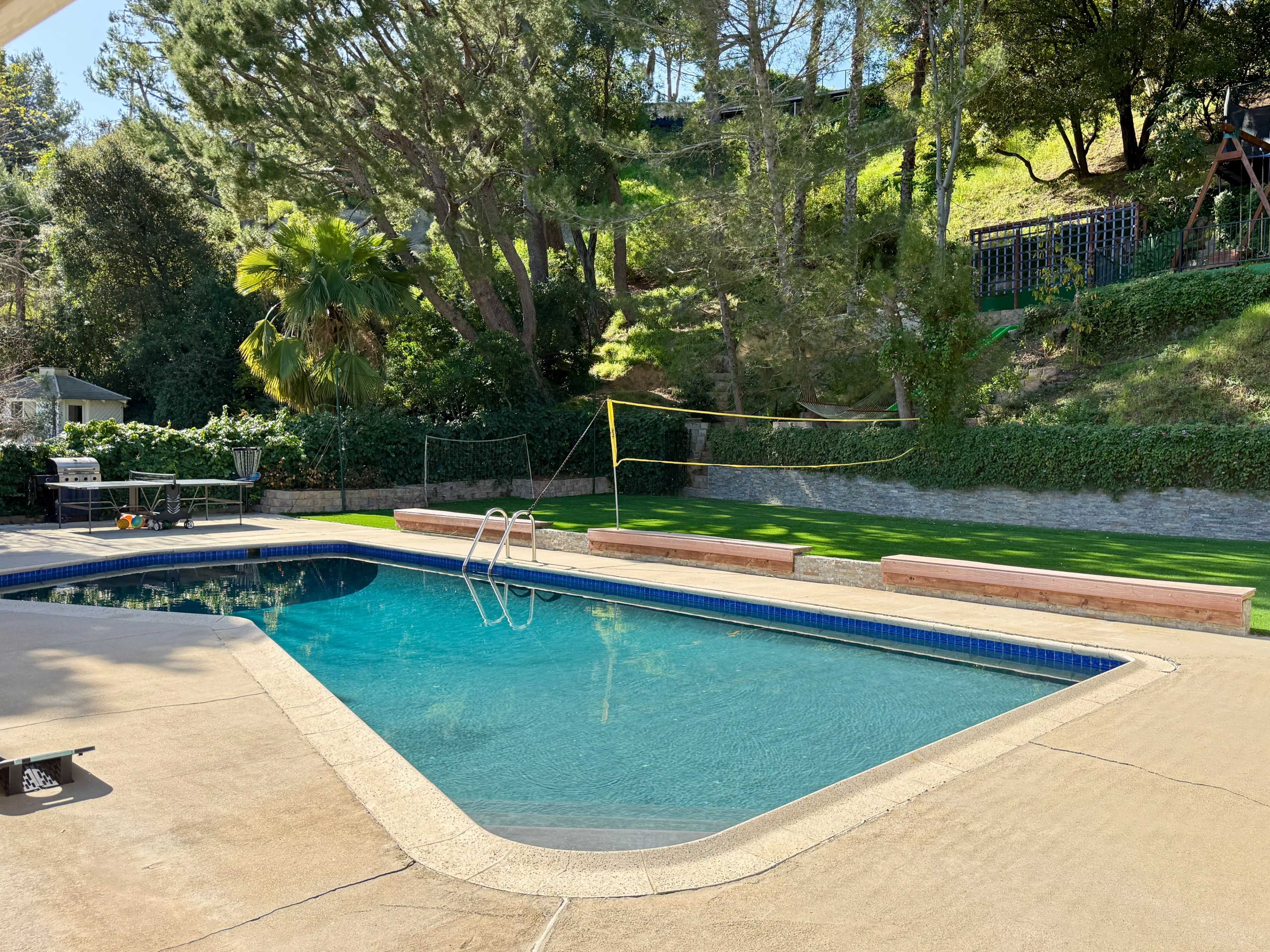 The image shows a swimming pool surrounded by a paved patio and lush greenery, with a volleyball net set up in the background.