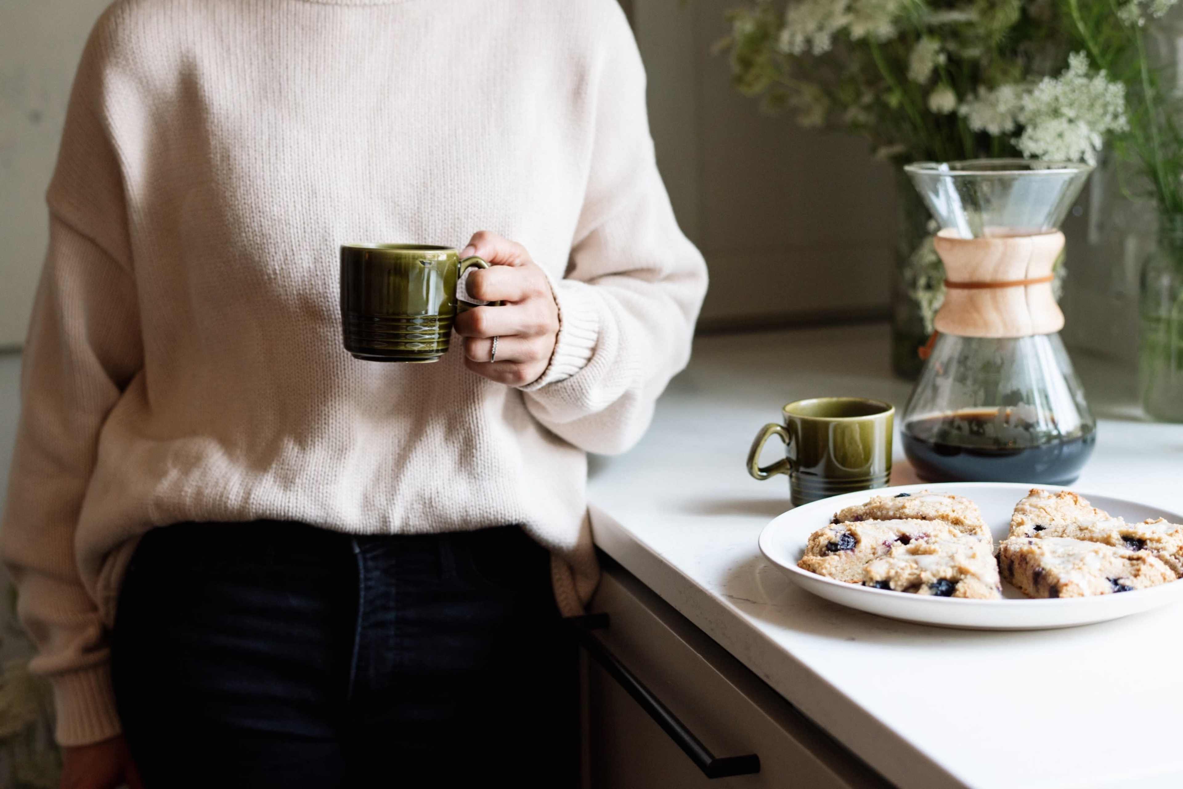 A person holding a green mug stands beside a plate of pastries and a coffee maker on a countertop.