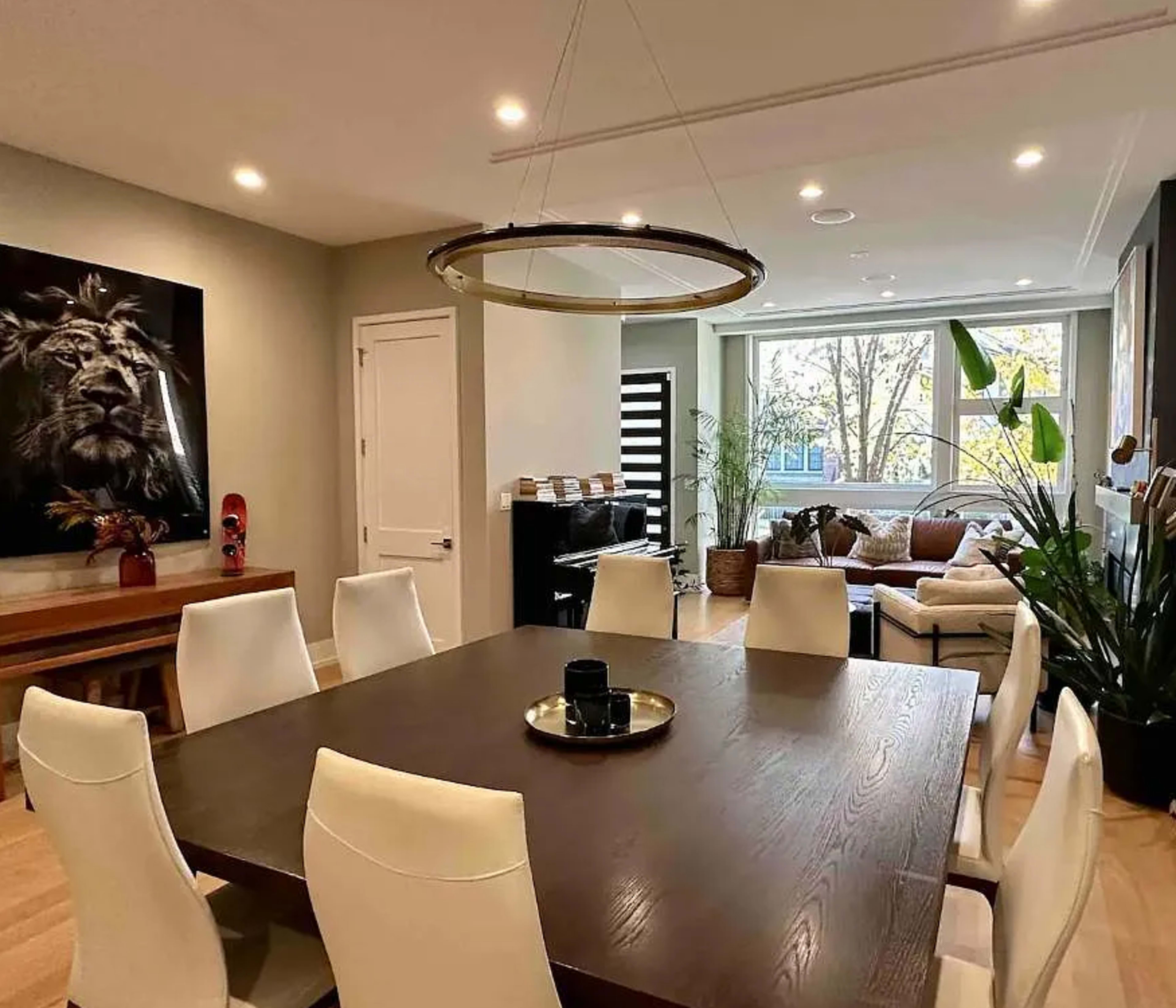 The image shows a modern dining room featuring a large wooden table surrounded by white chairs, with a lion artwork on the wall and a bright living area visible through a window.