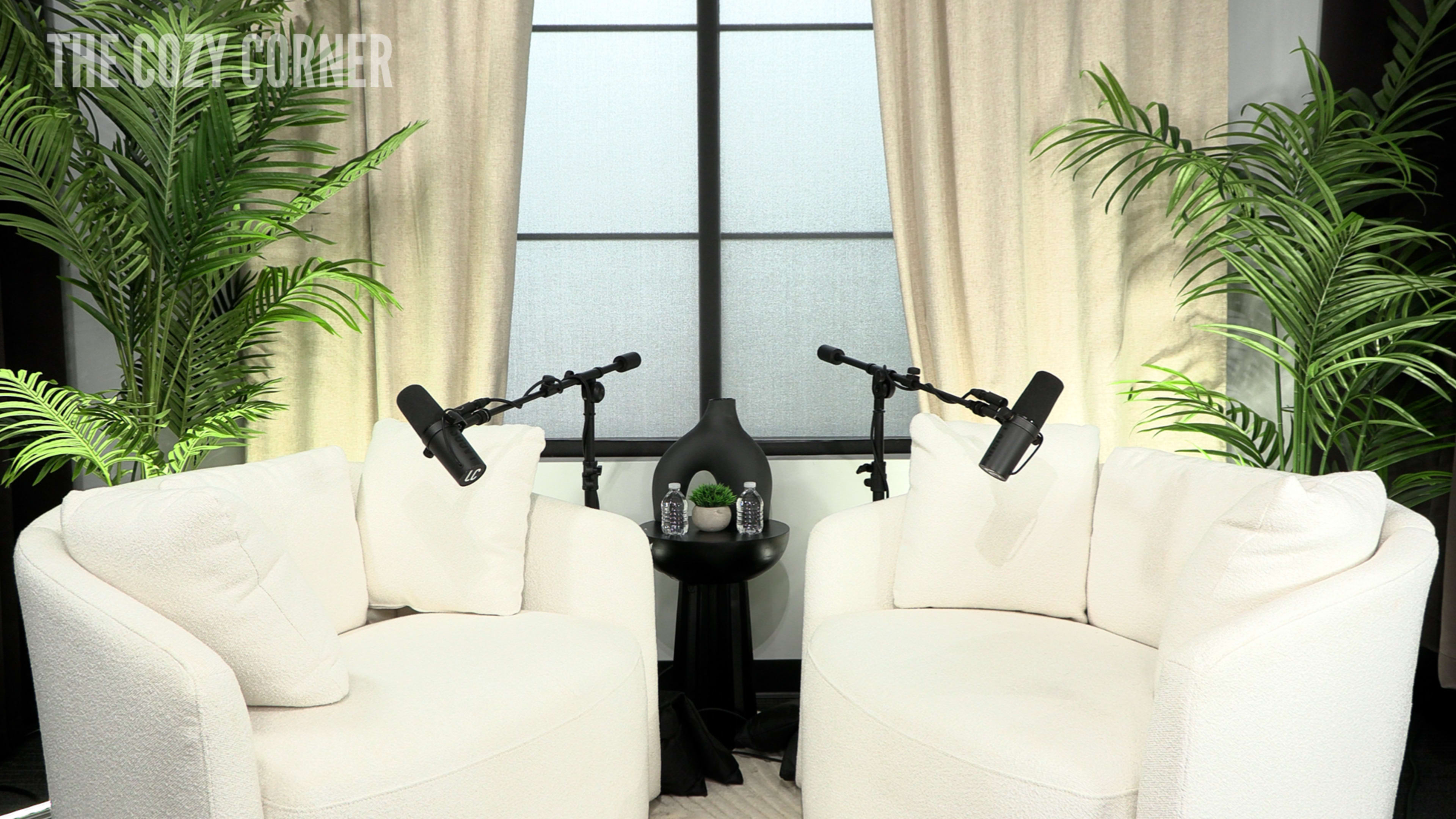 The image shows two white, upholstered chairs facing each other with a small black table in between, featuring two microphones and water bottles, all set against a backdrop of large curtains and greenery.