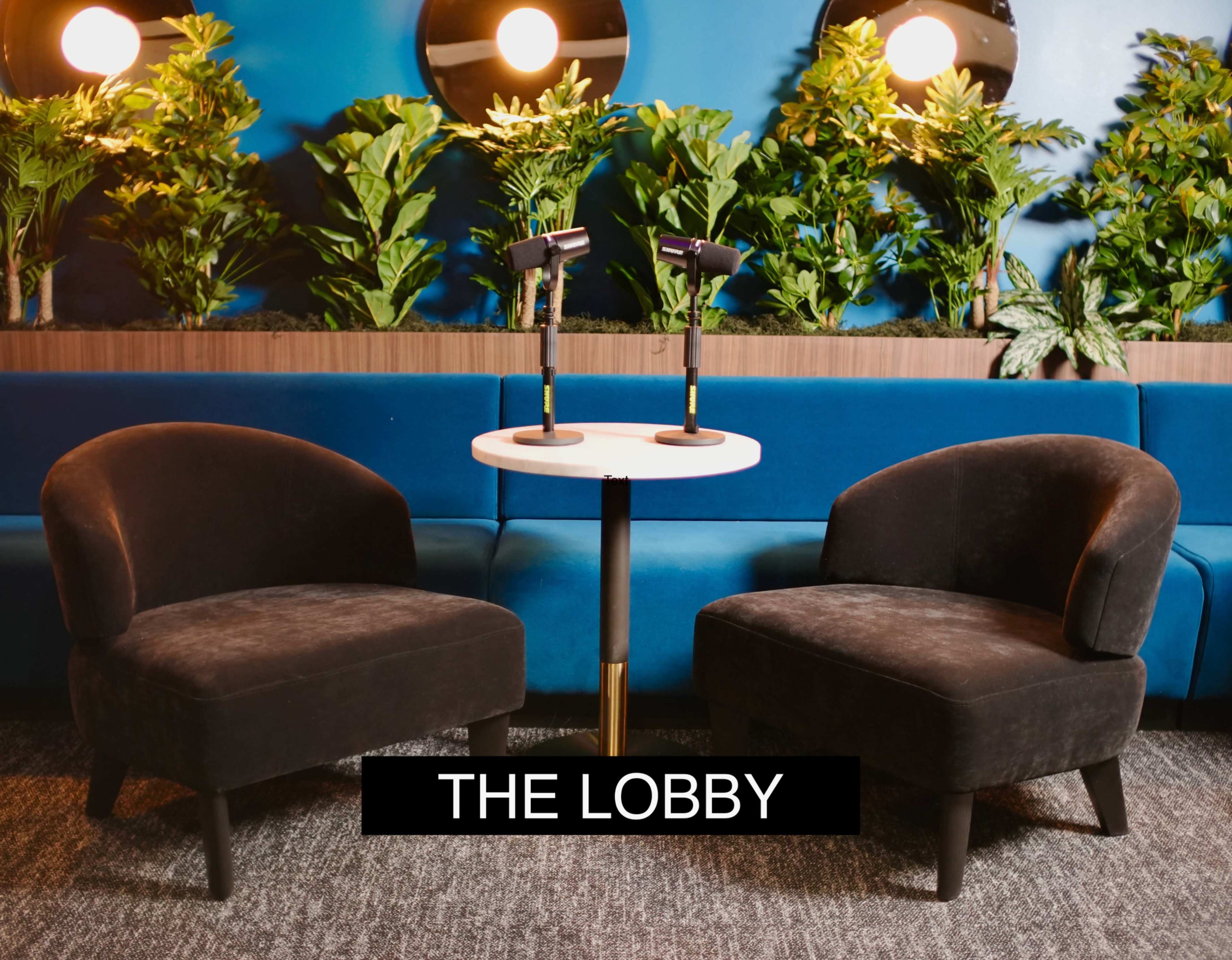 Two upholstered chairs are positioned around a small round table in a lobby area, accompanied by decorative plants on the walls.