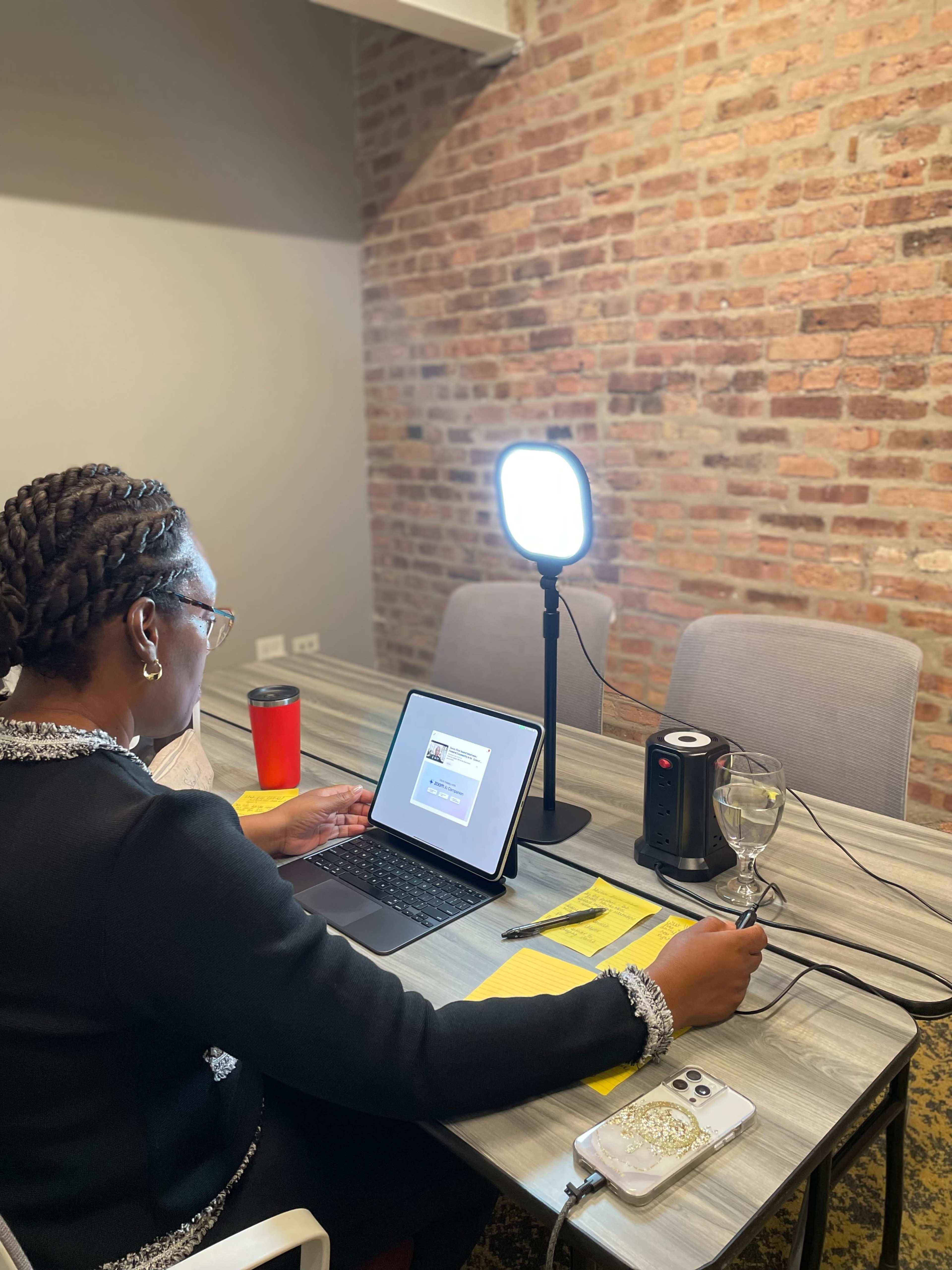 A person is sitting at a table with a laptop, a lighting setup, and various items, including a glass of wine and a red travel mug, against a backdrop of a brick wall.