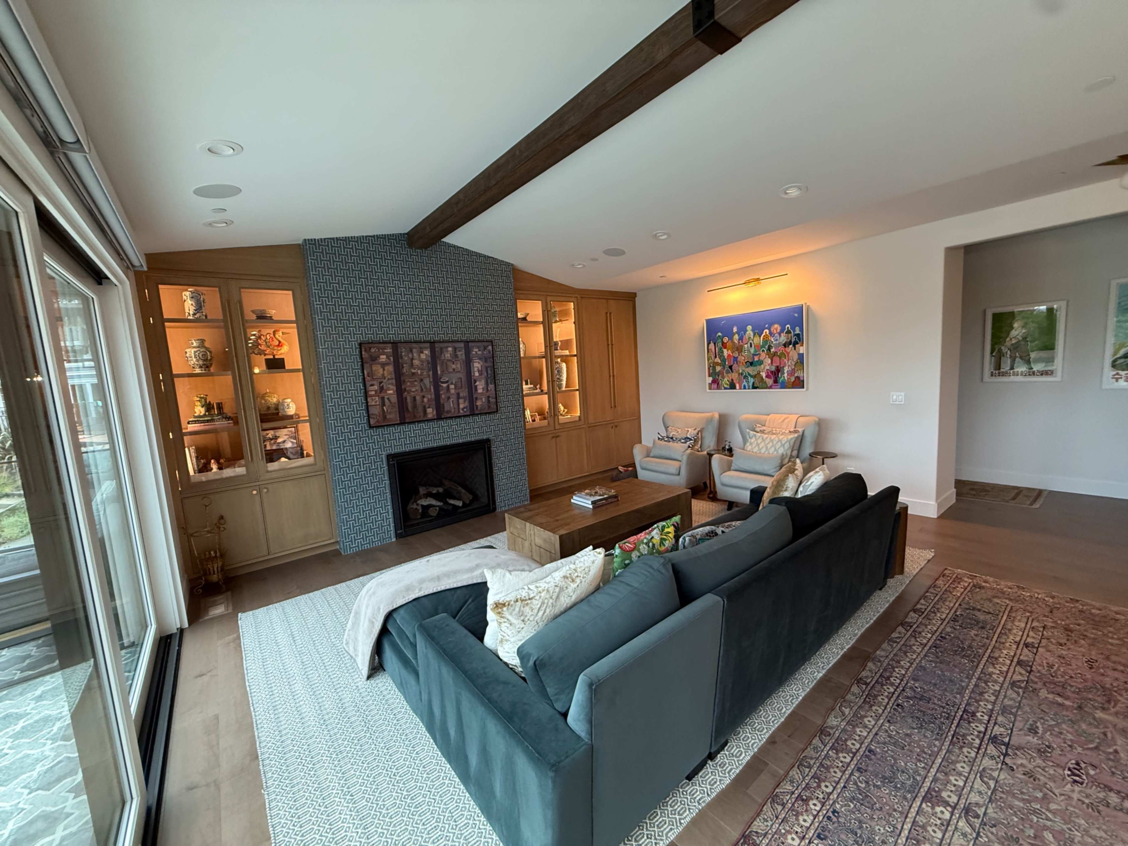A modern living room features a couch facing a fireplace, flanked by built-in cabinetry with decorative items and a large window showcasing an outdoor view.