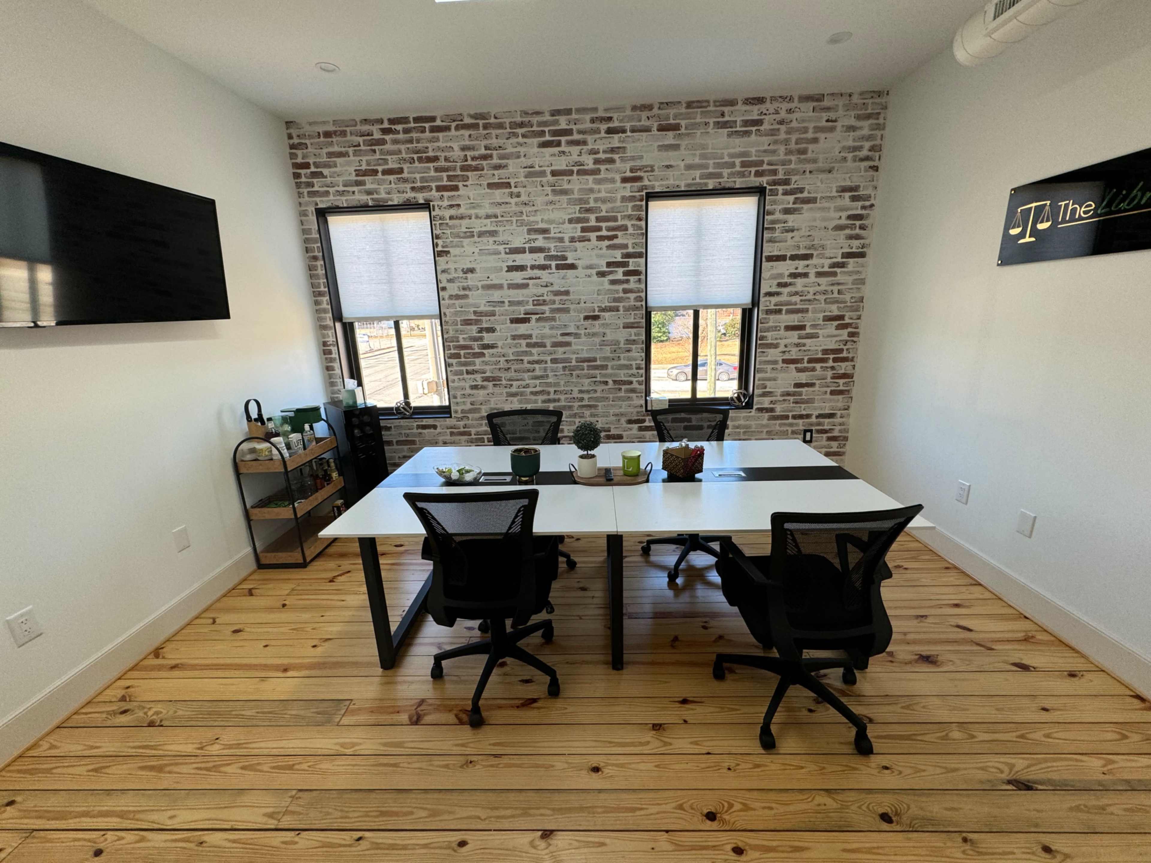 A modern office space with a white table, black chairs, and a brick accent wall, featuring two windows and a small storage unit.