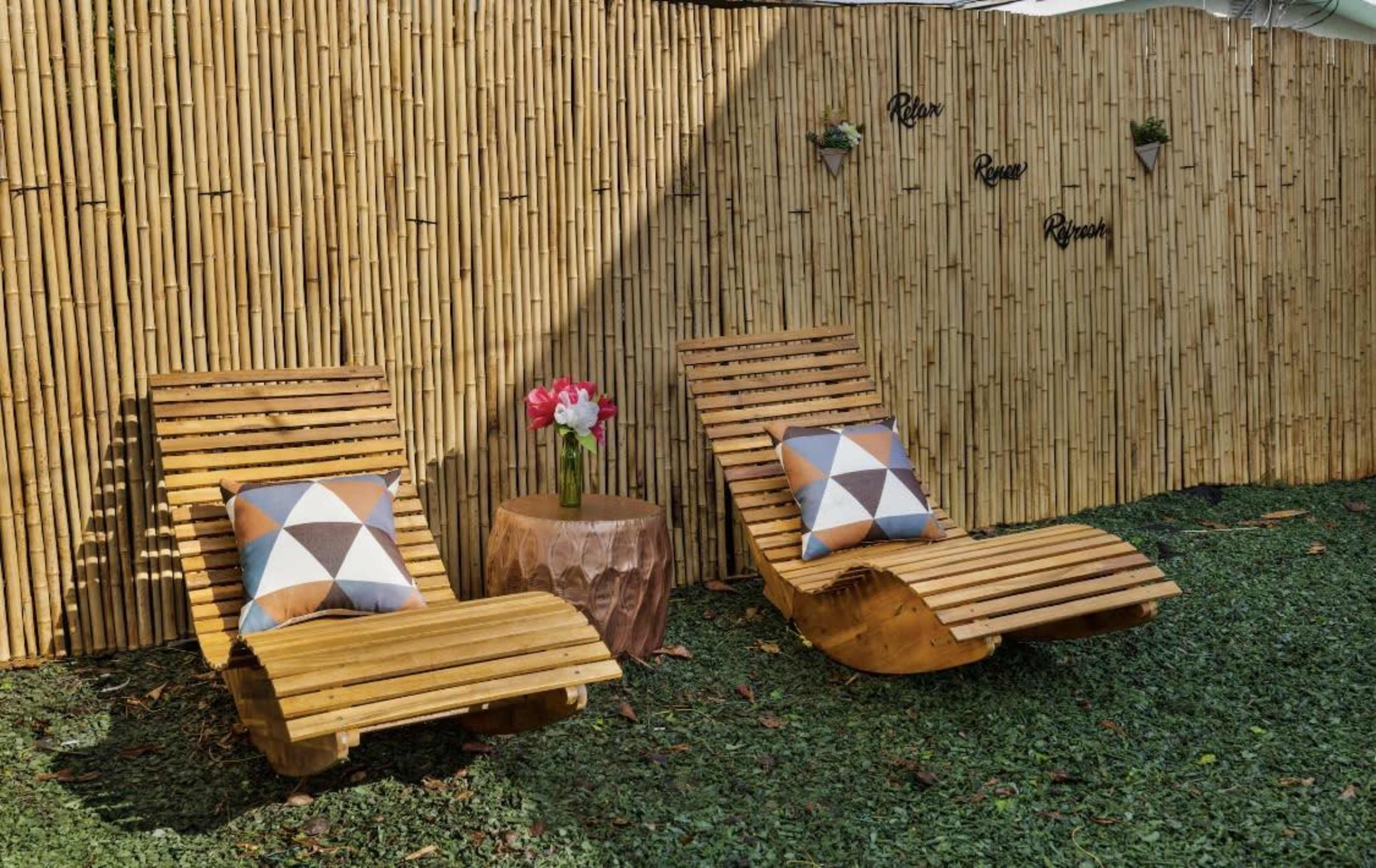 Two wooden lounge chairs with patterned cushions beside a small wooden table and a bamboo fence backdrop.