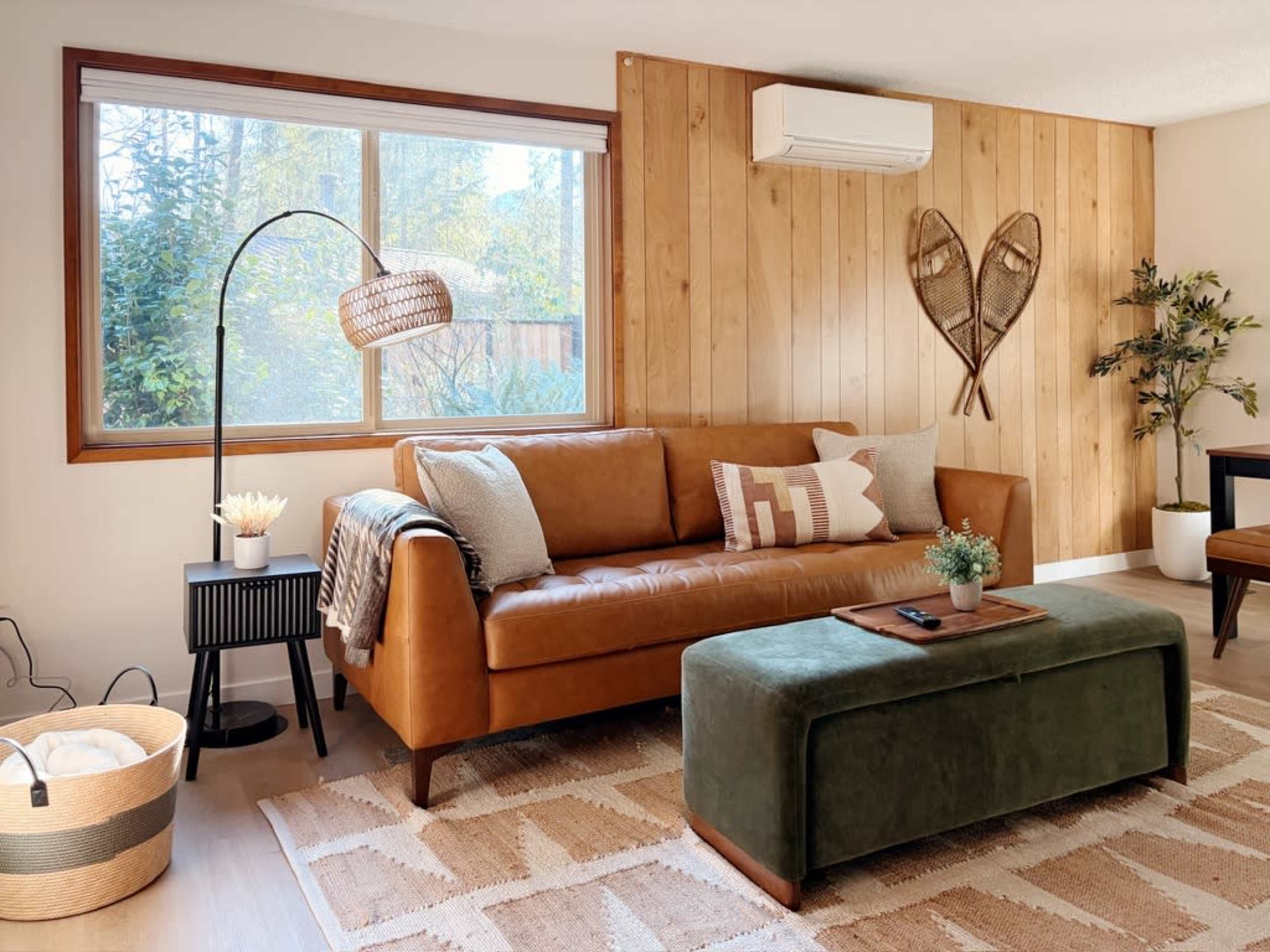 The image shows a cozy living room with a brown leather sofa, a green ottoman, and a window that lets in natural light, complemented by wooden wall paneling.