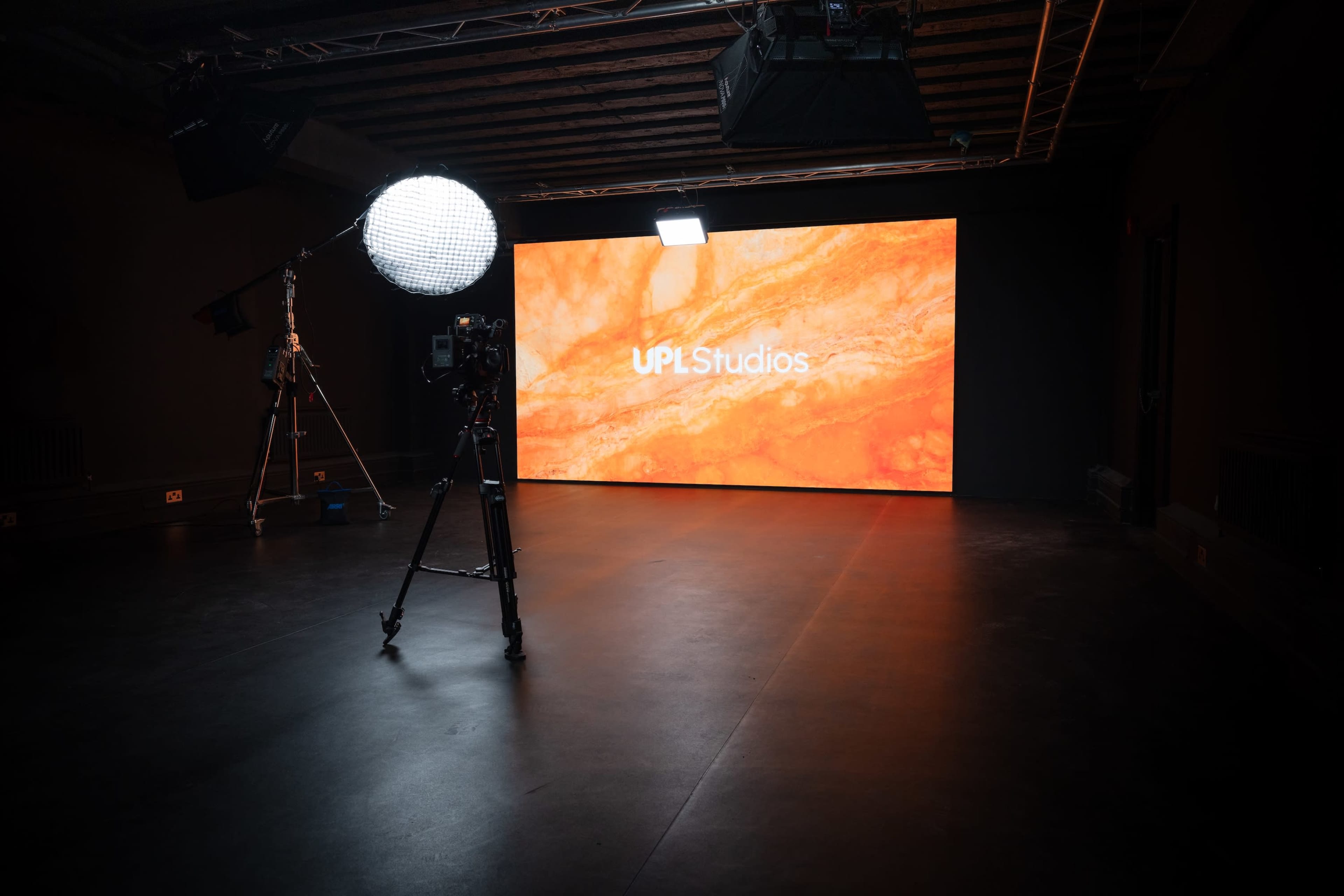 A photography studio setup features a large illuminated screen displaying "UPL Studios," with cameras and lighting equipment positioned on the floor.